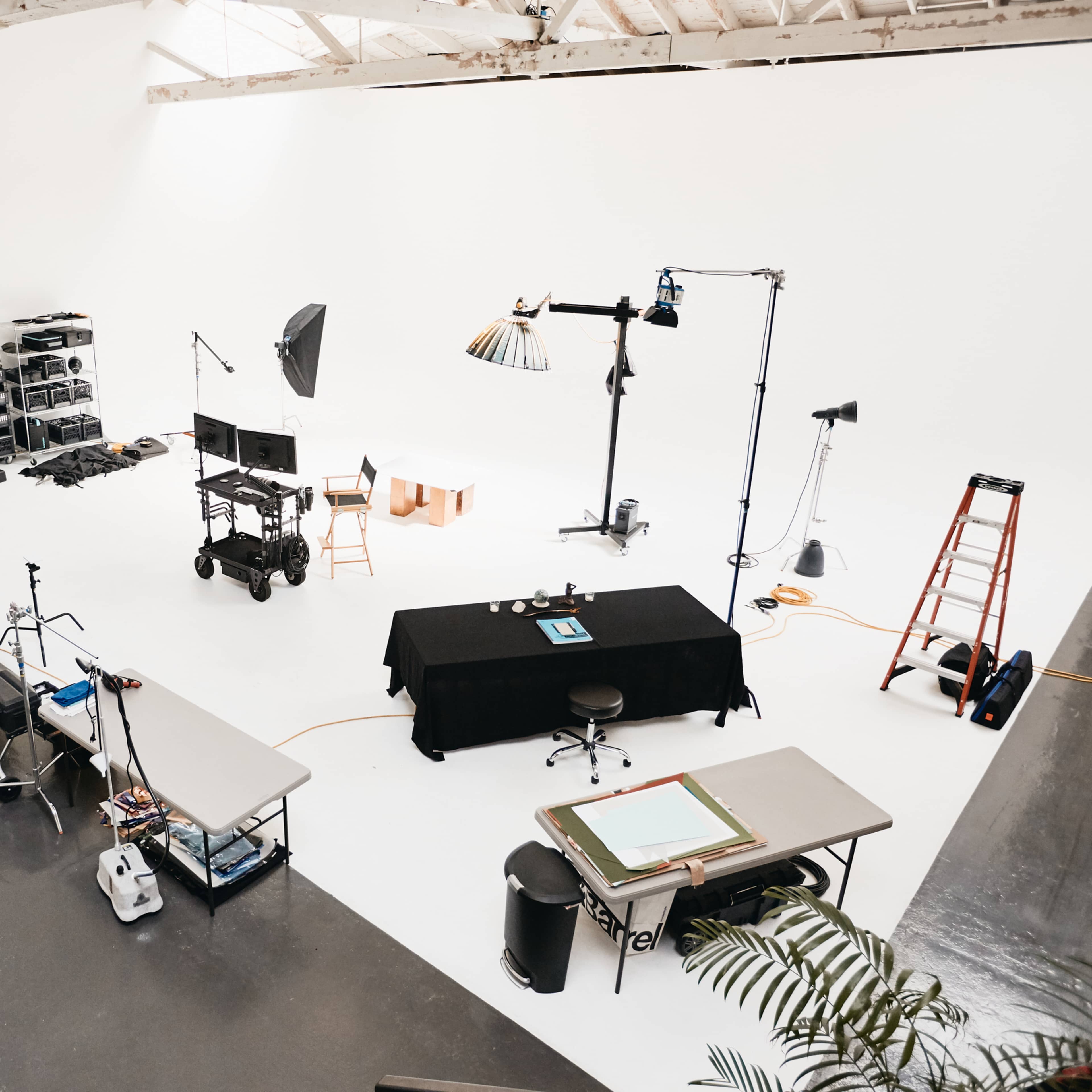 A spacious photography studio is set up with various lighting equipment, tables, and a ladder against a plain white backdrop.