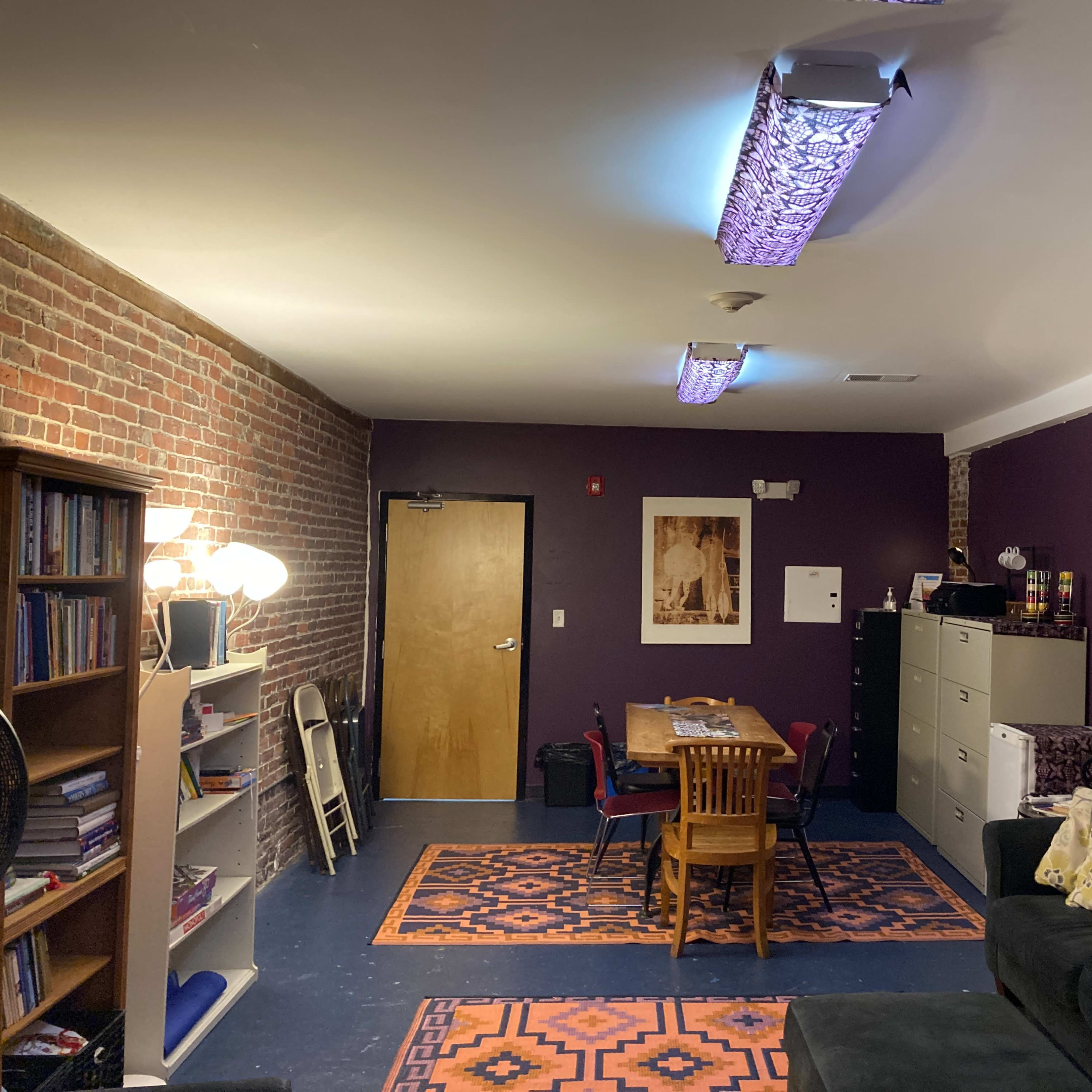 A room with purple walls, a brick wall section, a table with chairs, a couch, and shelving filled with books.