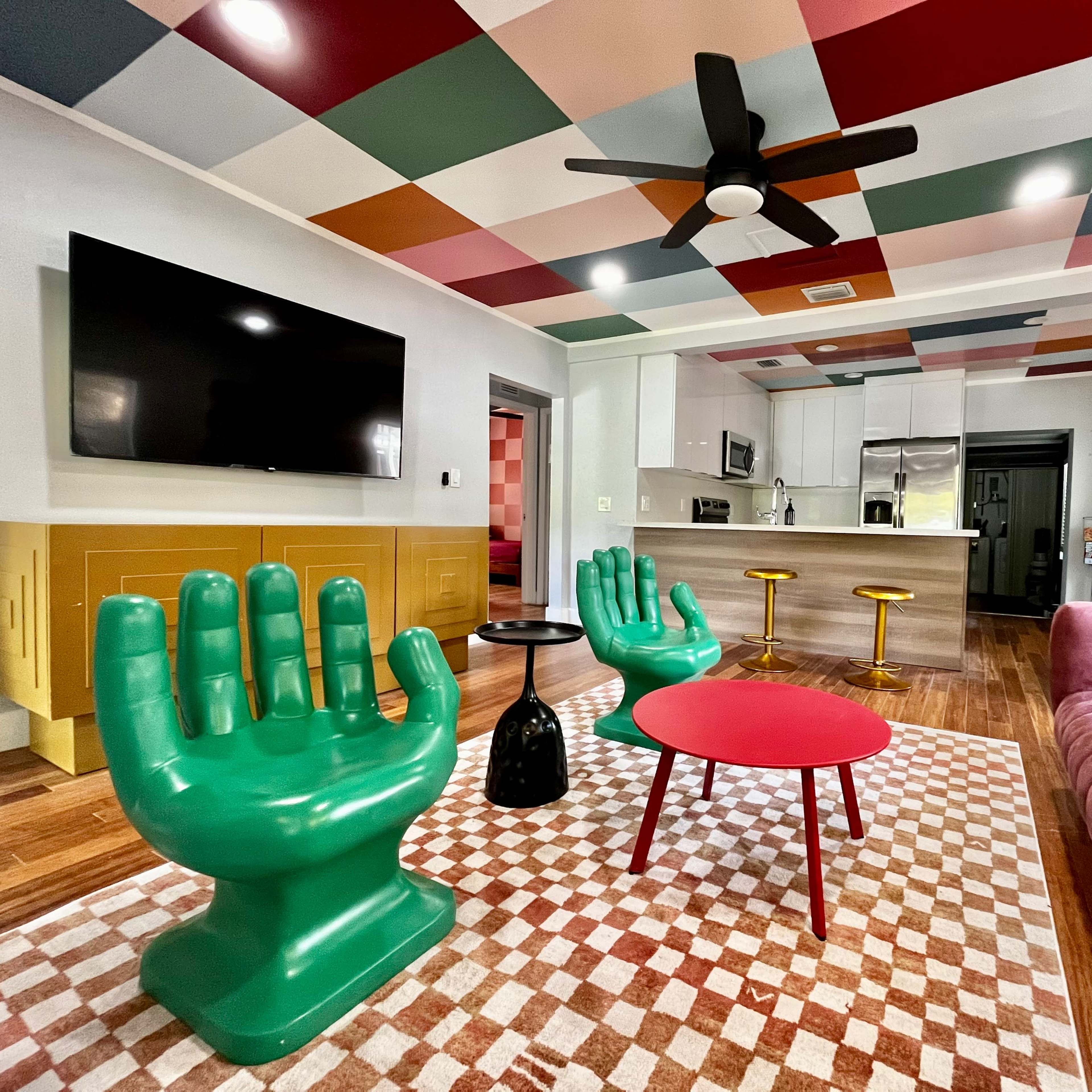 A vibrant living room featuring bright green hand-shaped chairs, a pink round coffee table, and a sofa, with a colorful patterned ceiling and an open kitchen in the background.