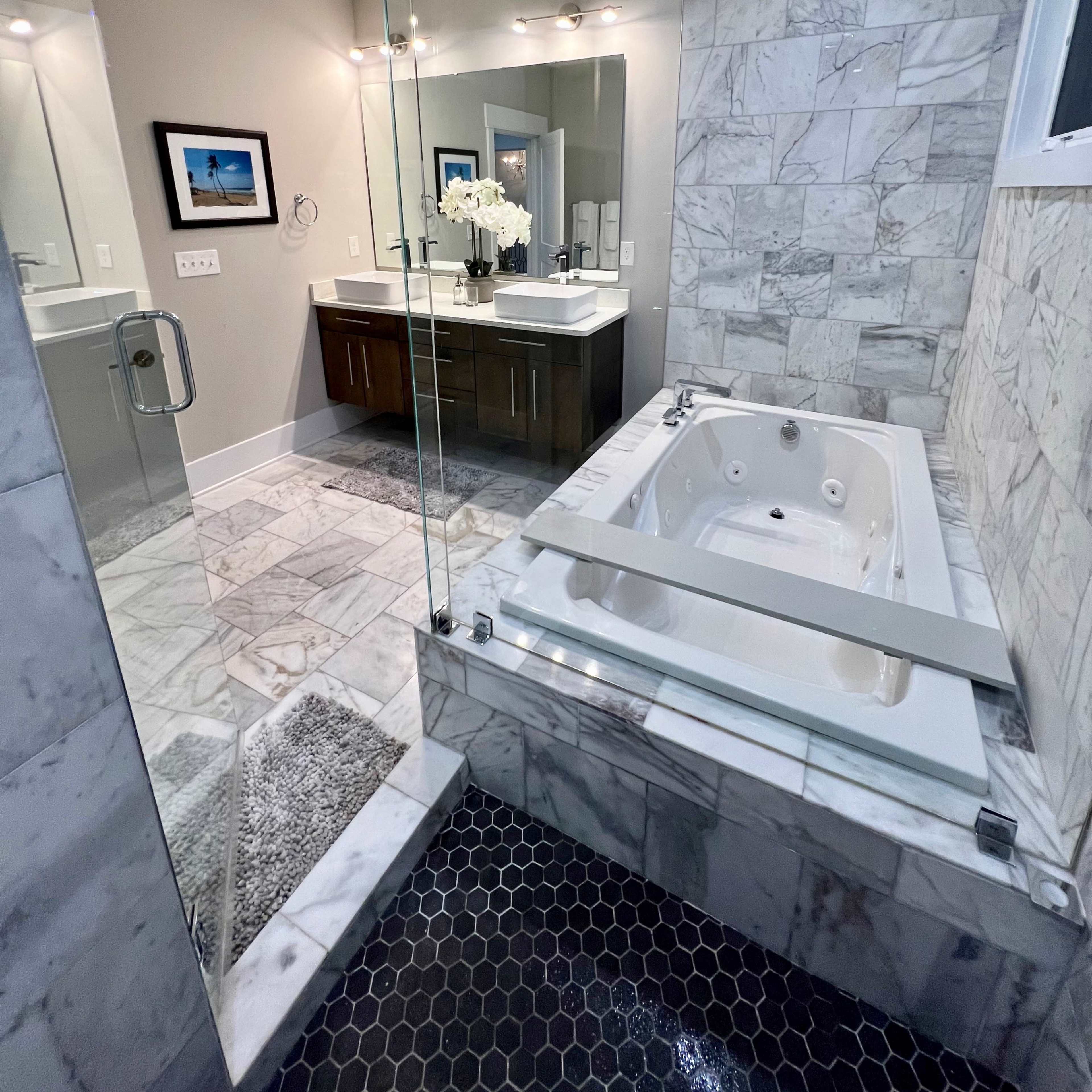 A modern bathroom featuring a glass shower enclosure, a freestanding bathtub with jets, and a double vanity with a large mirror and decorative flower arrangement.