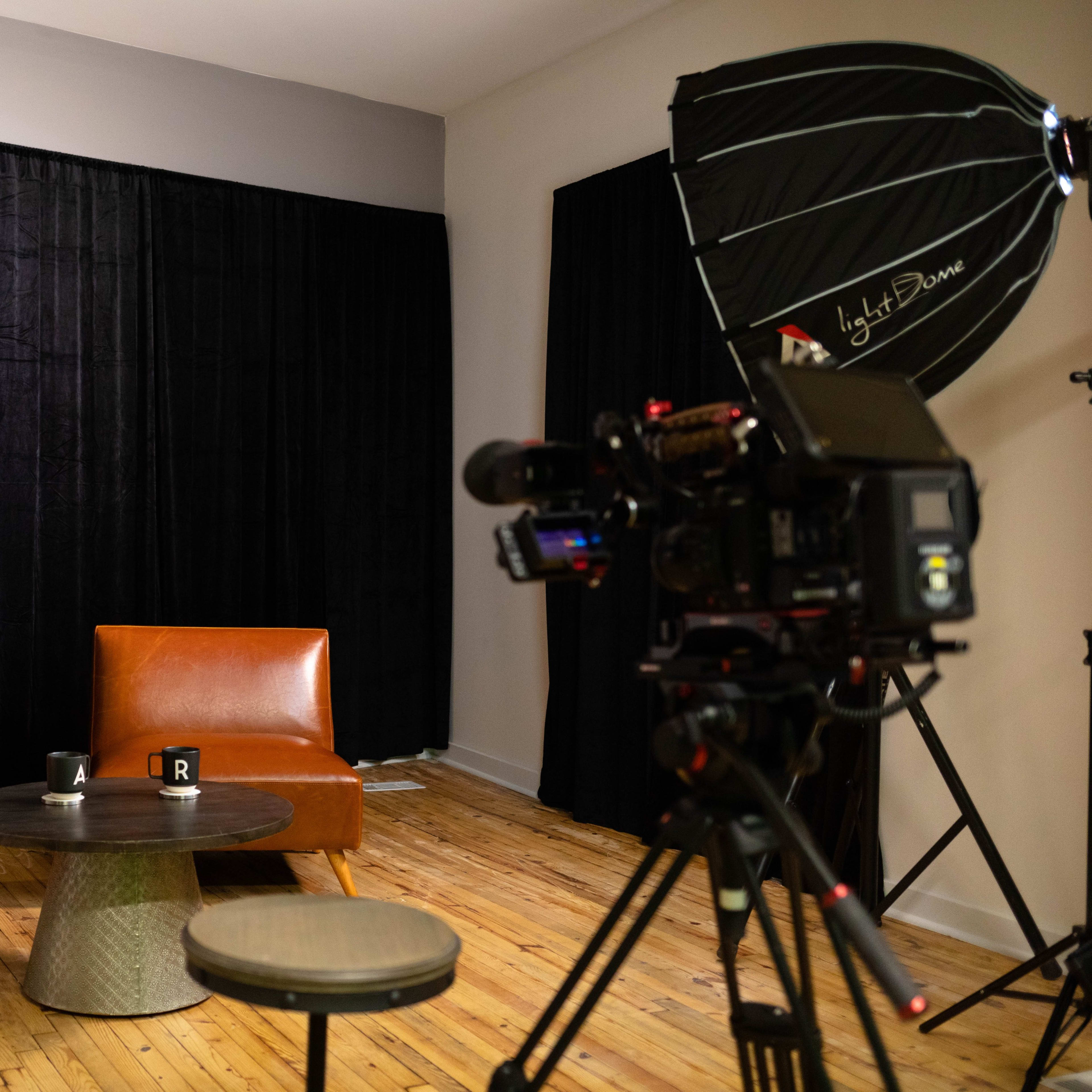 The scene features two brown chairs and a round table set against a black backdrop, with a camera on a tripod positioned to the right.