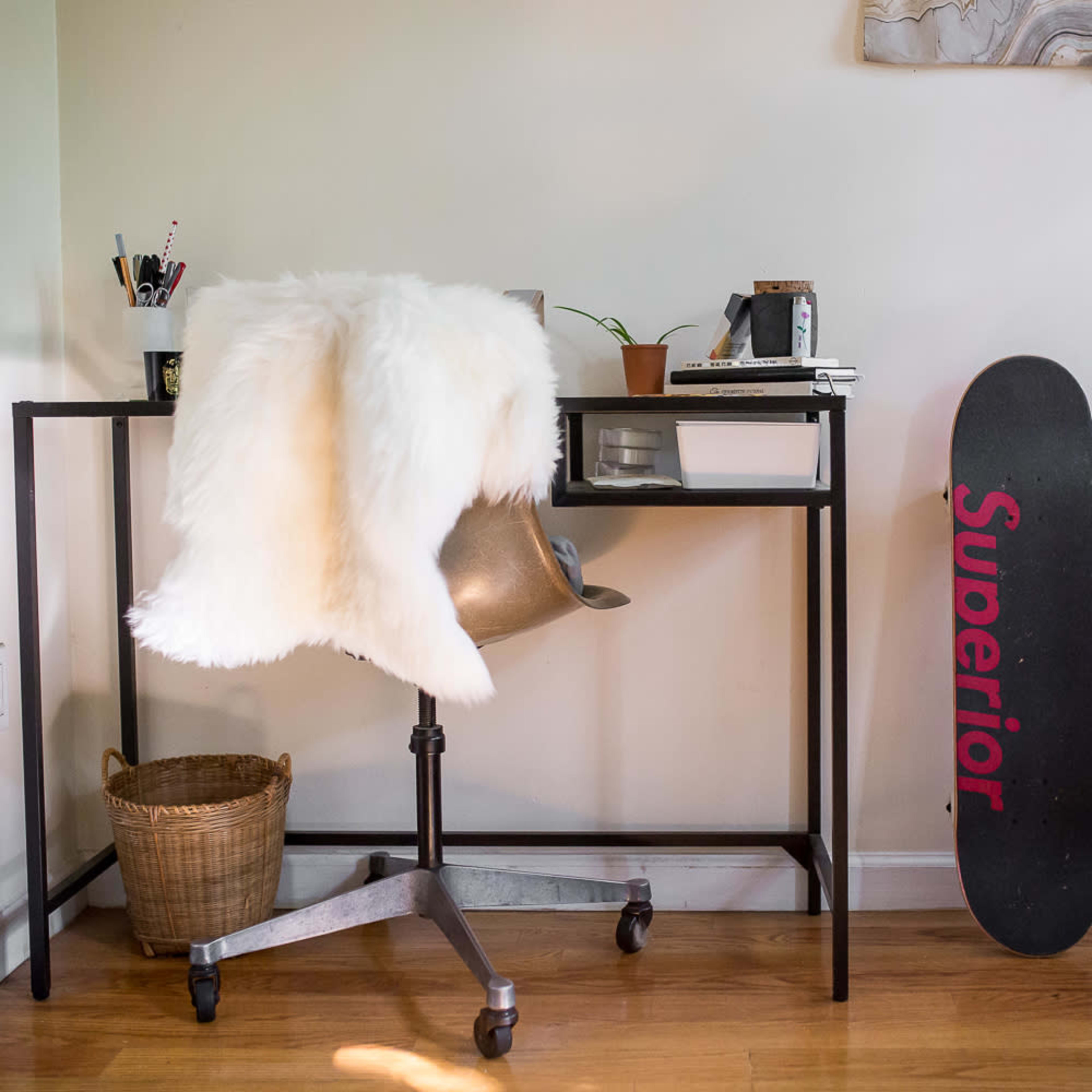 A modern workspace features a black metal desk with an ergonomic chair, a fur throw, a potted plant, and a skateboard propped against the wall.