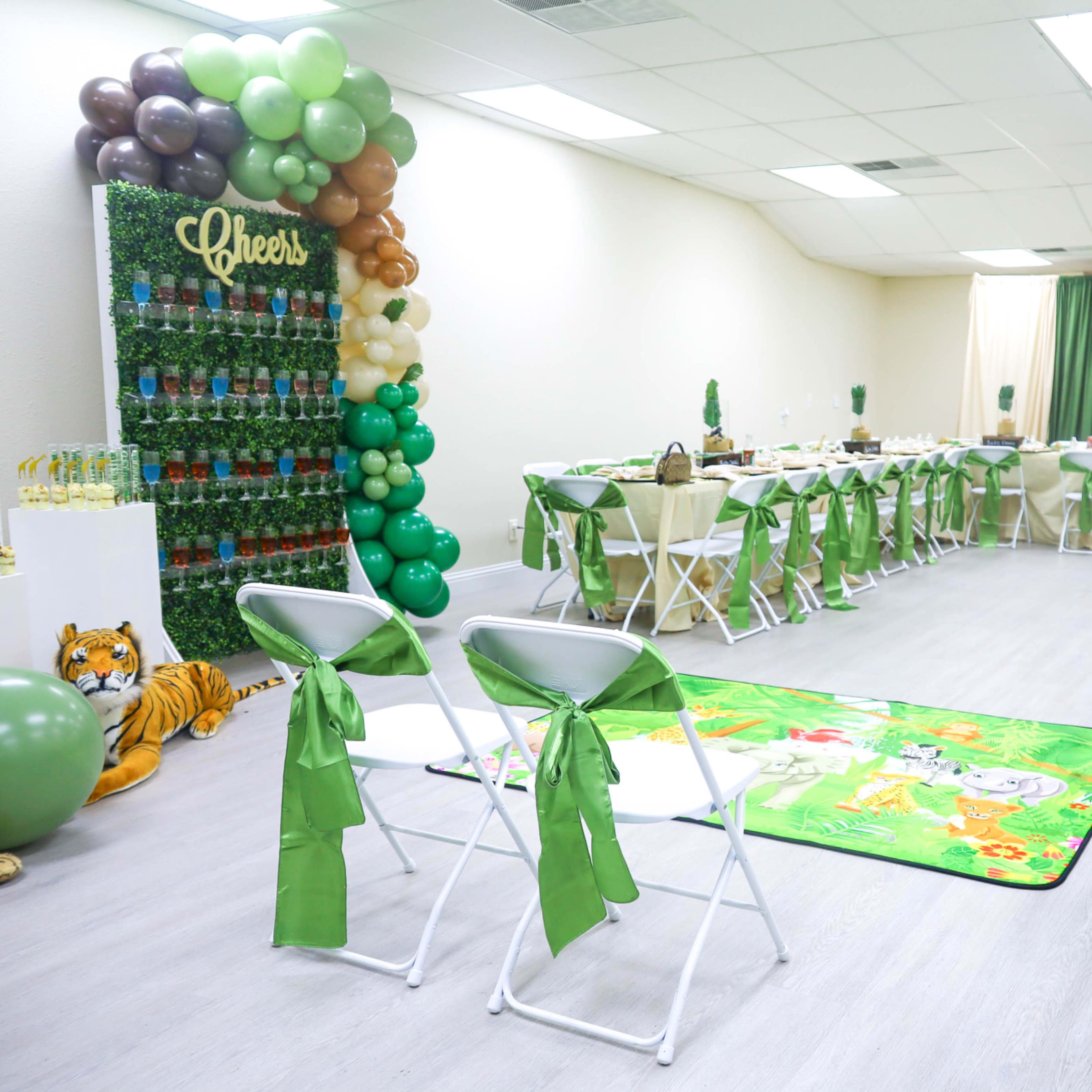 A decorated event space with tables set for a gathering, featuring green and brown balloons, floral arrangements, and a themed jungle rug.