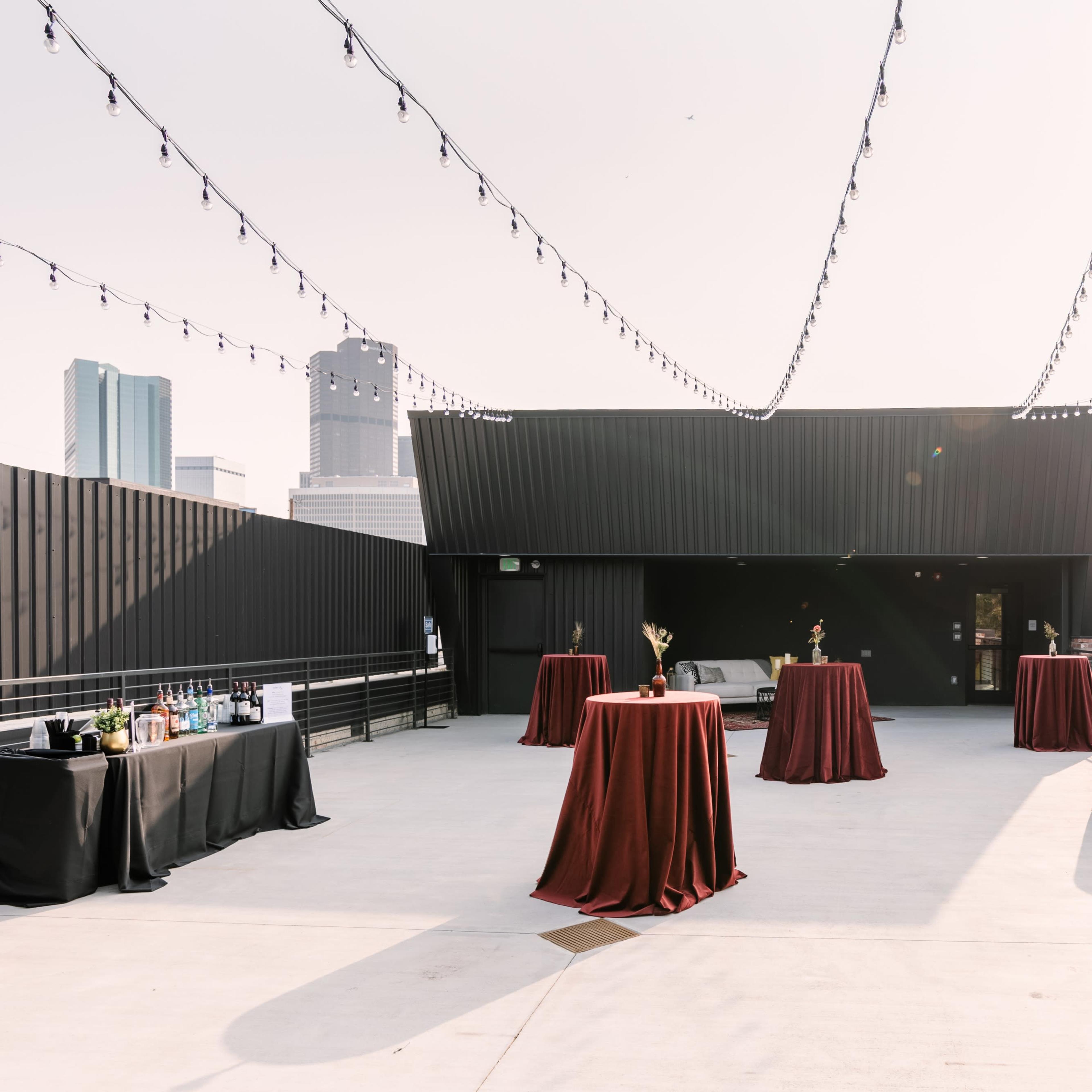 The scene depicts a rooftop event space with several round tables covered in maroon tablecloths, string lights overhead, and a city skyline in the background.