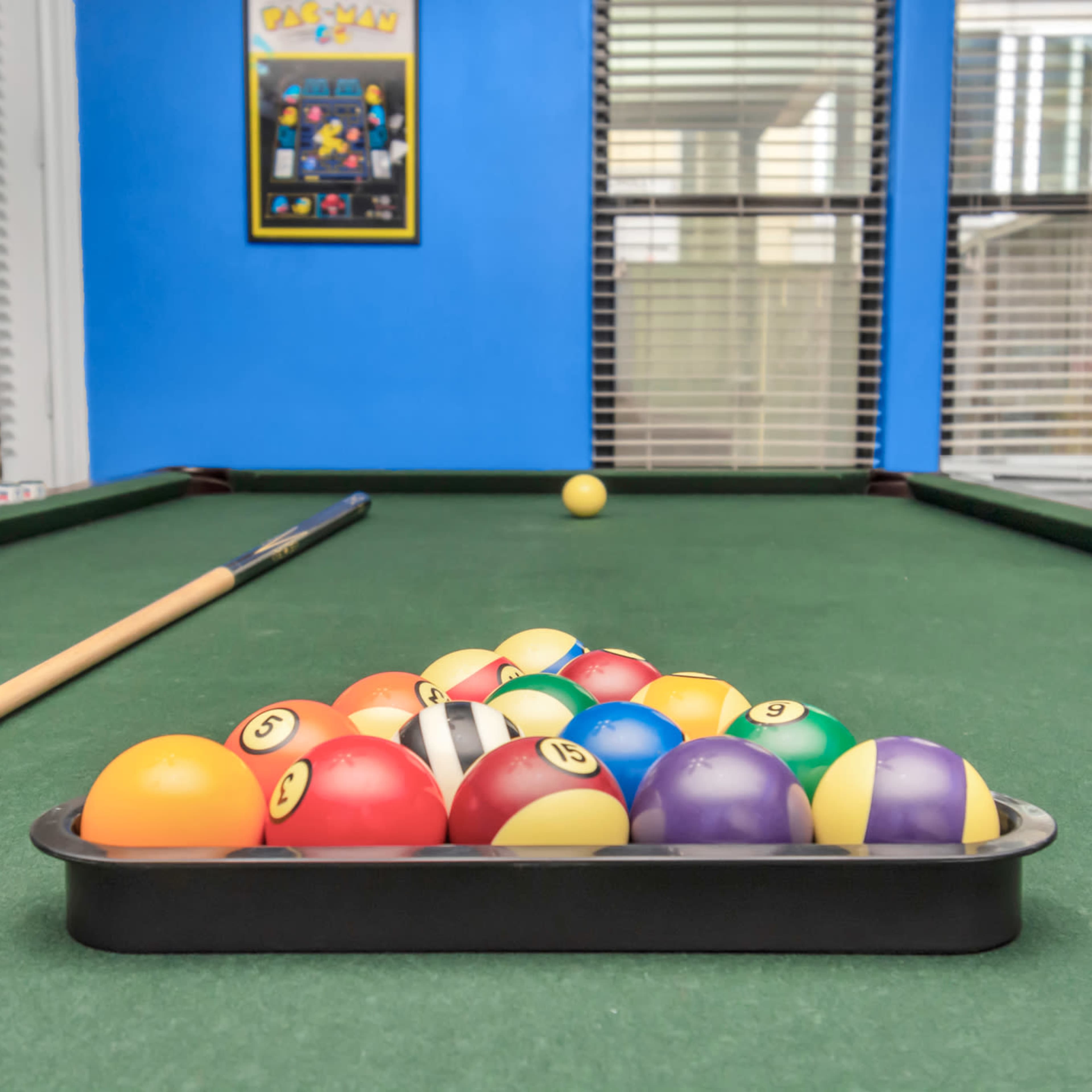 A set of colorful pool balls is arranged in a tray on a green billiard table, with a cue stick resting beside it and a single white ball visible in the background.