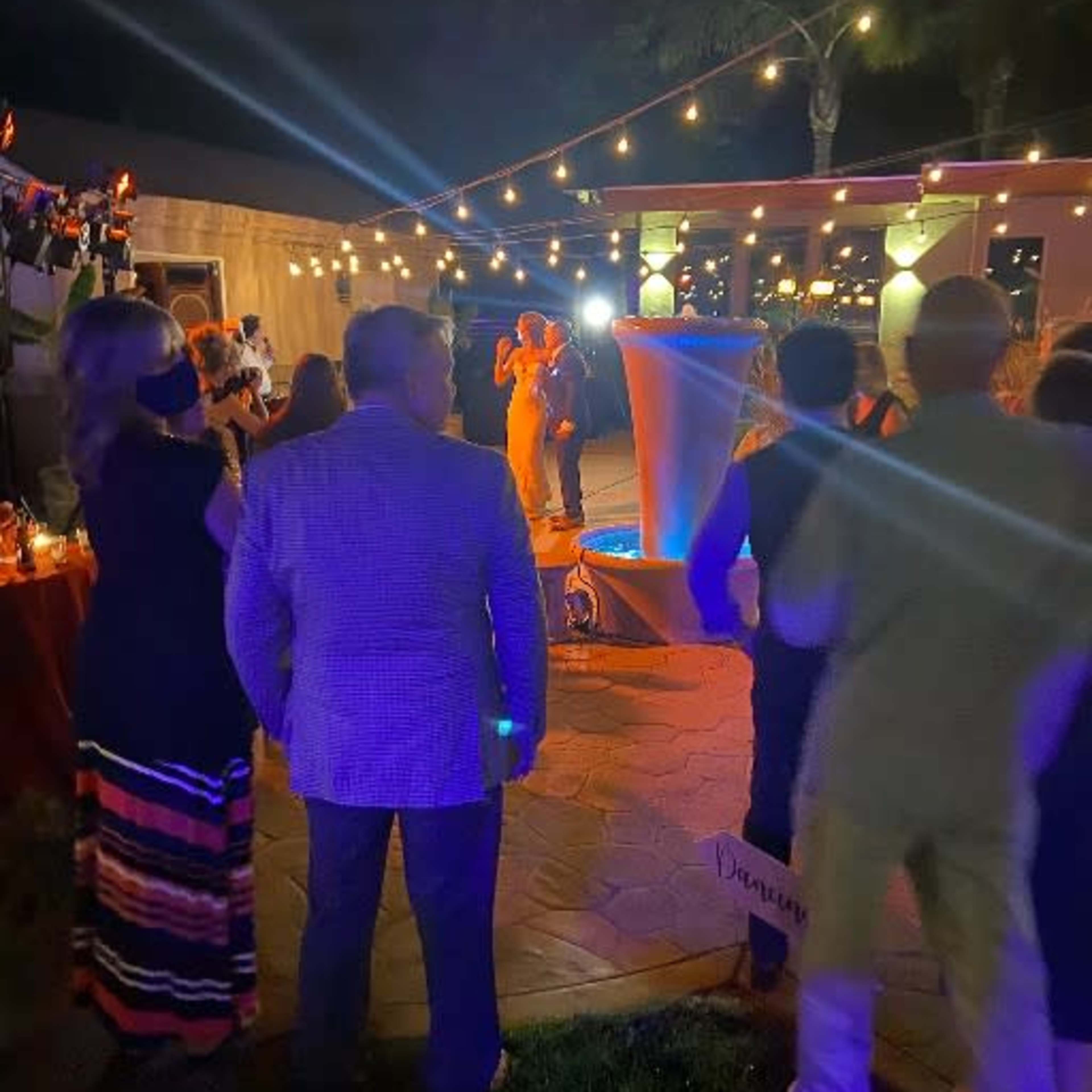 A group of people gathers in an outdoor setting at night, with string lights illuminating the scene as a couple dances in front of them.