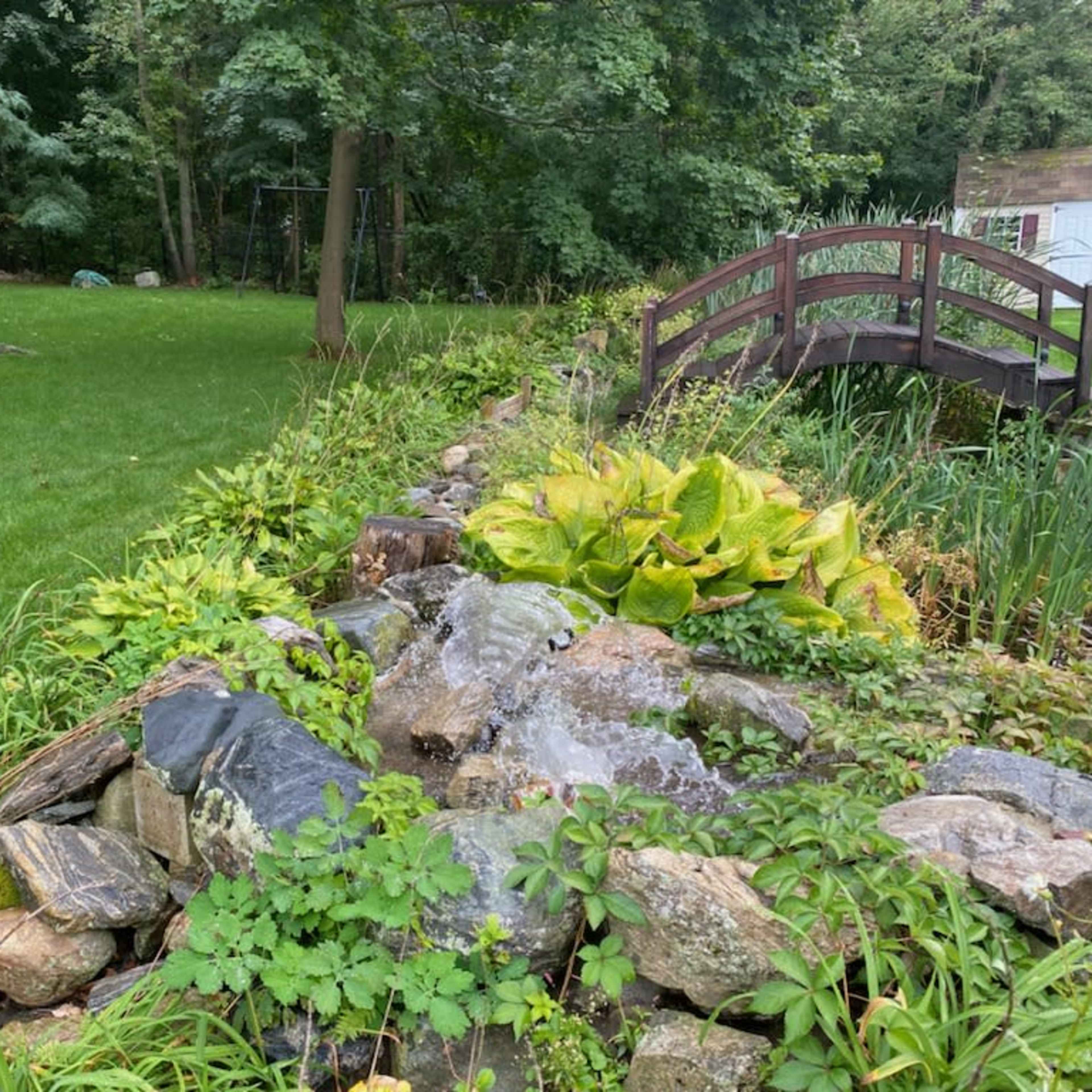 A small waterfall cascades over rocks beside a lush green lawn and wooden bridge in a landscaped garden.