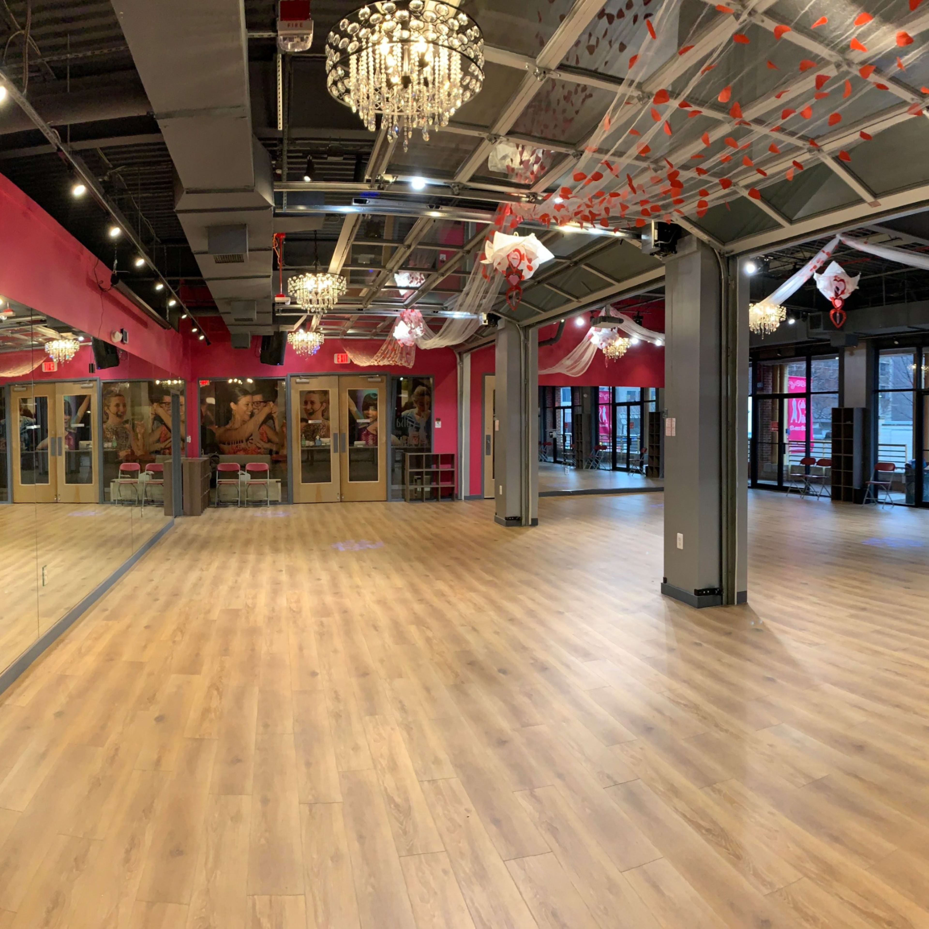 A spacious dance studio features wooden flooring, large mirrors along one wall, and multiple windows providing natural light.