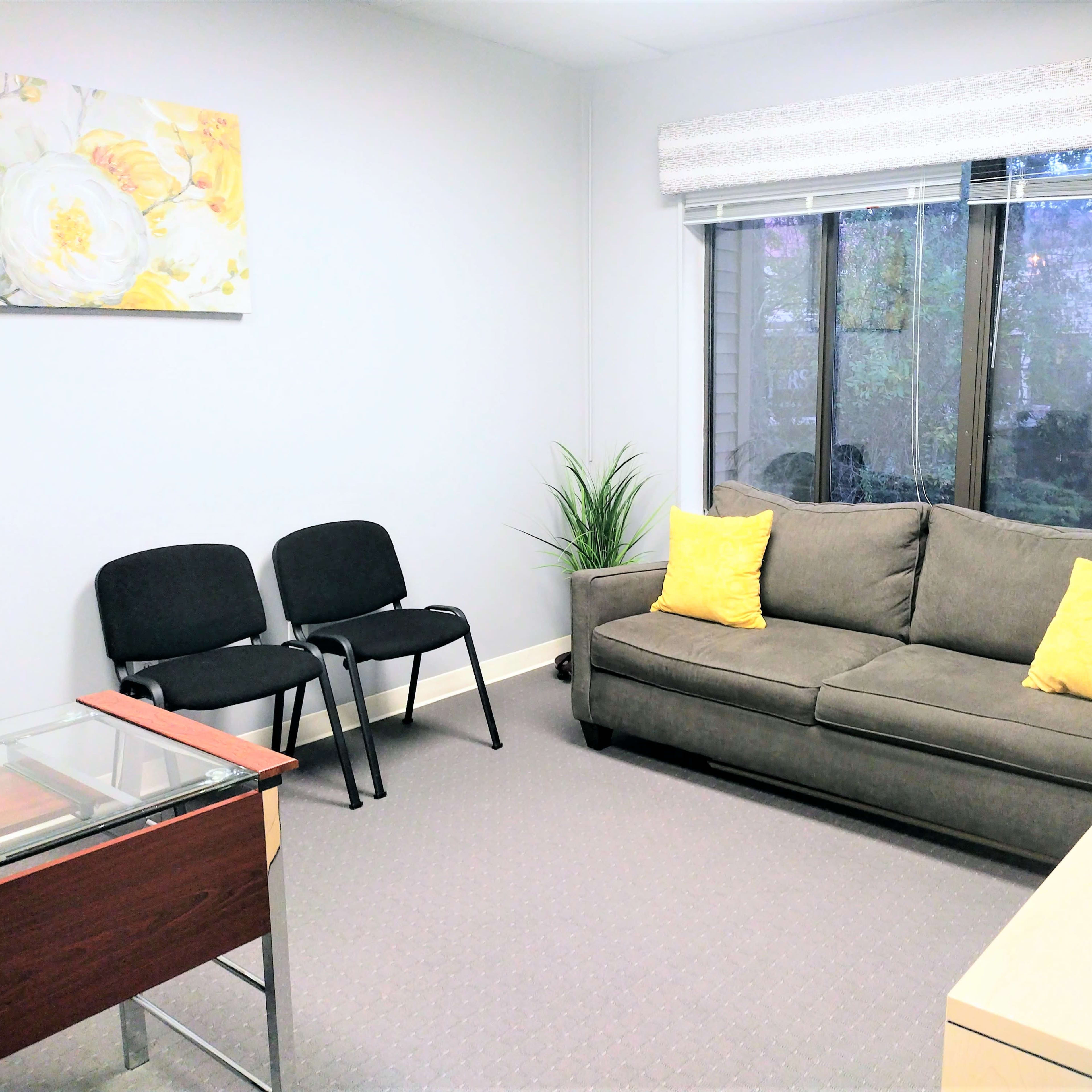 A small office with a sofa, two chairs, a glass table, and a window overlooking greenery.
