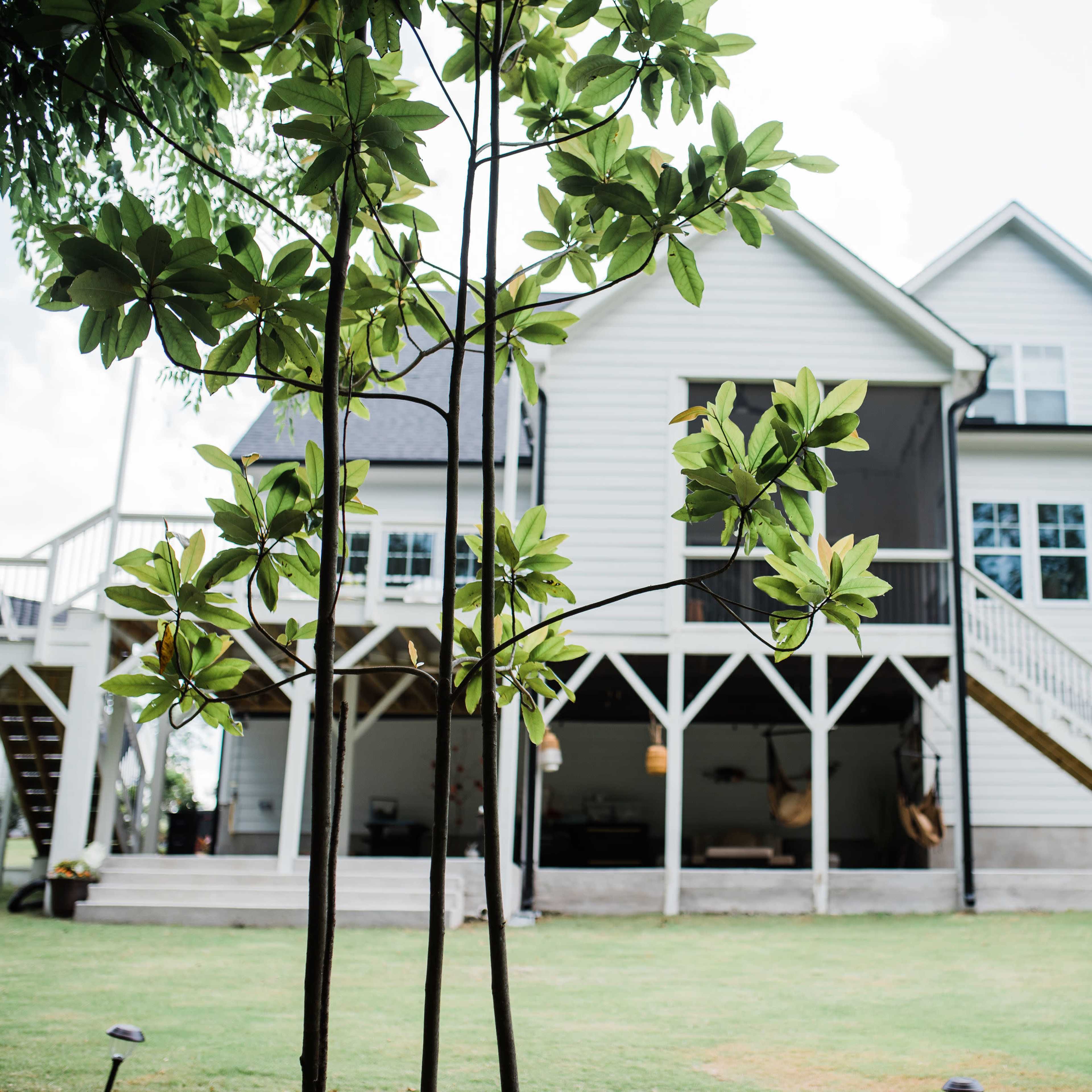 A two-story house with a wraparound porch and multiple windows is surrounded by green grass and trees.