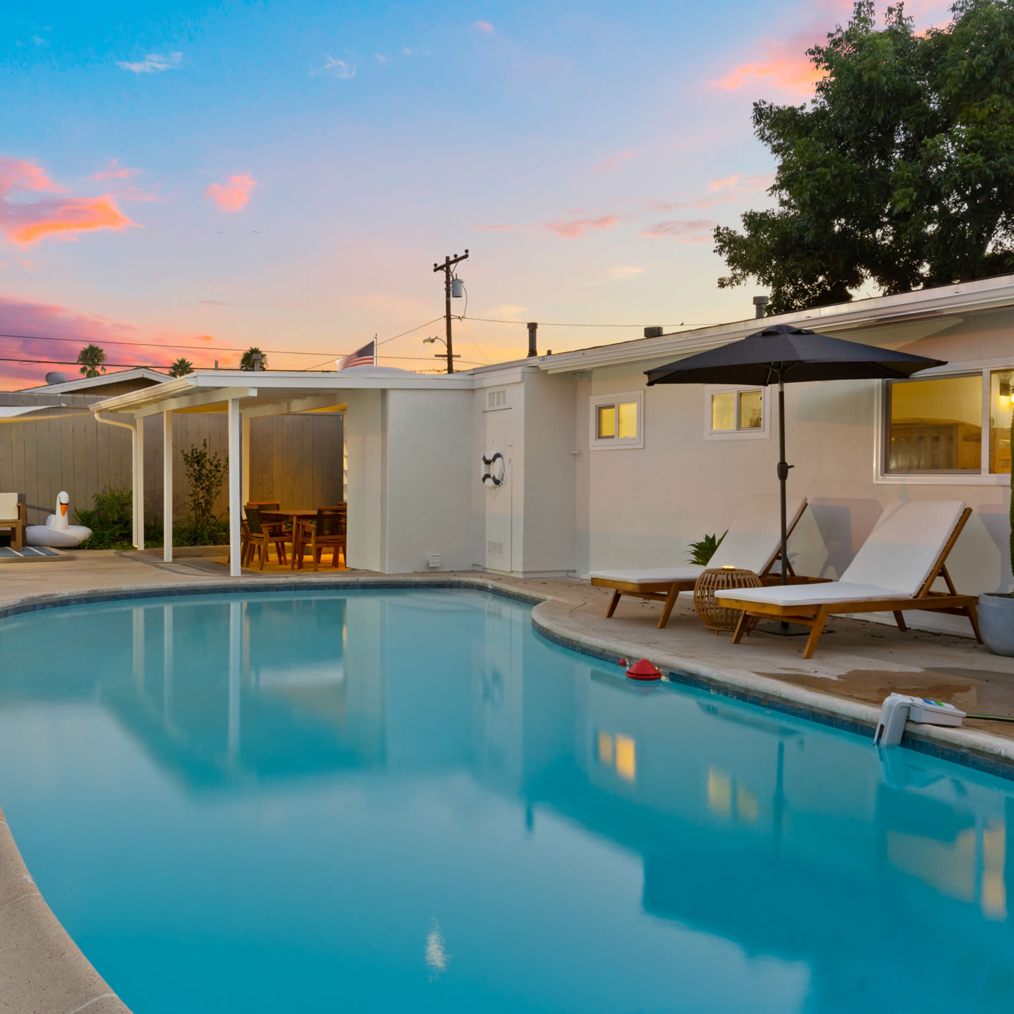 A swimming pool area surrounded by lounge chairs and plants, with a pink and blue sky at sunset.