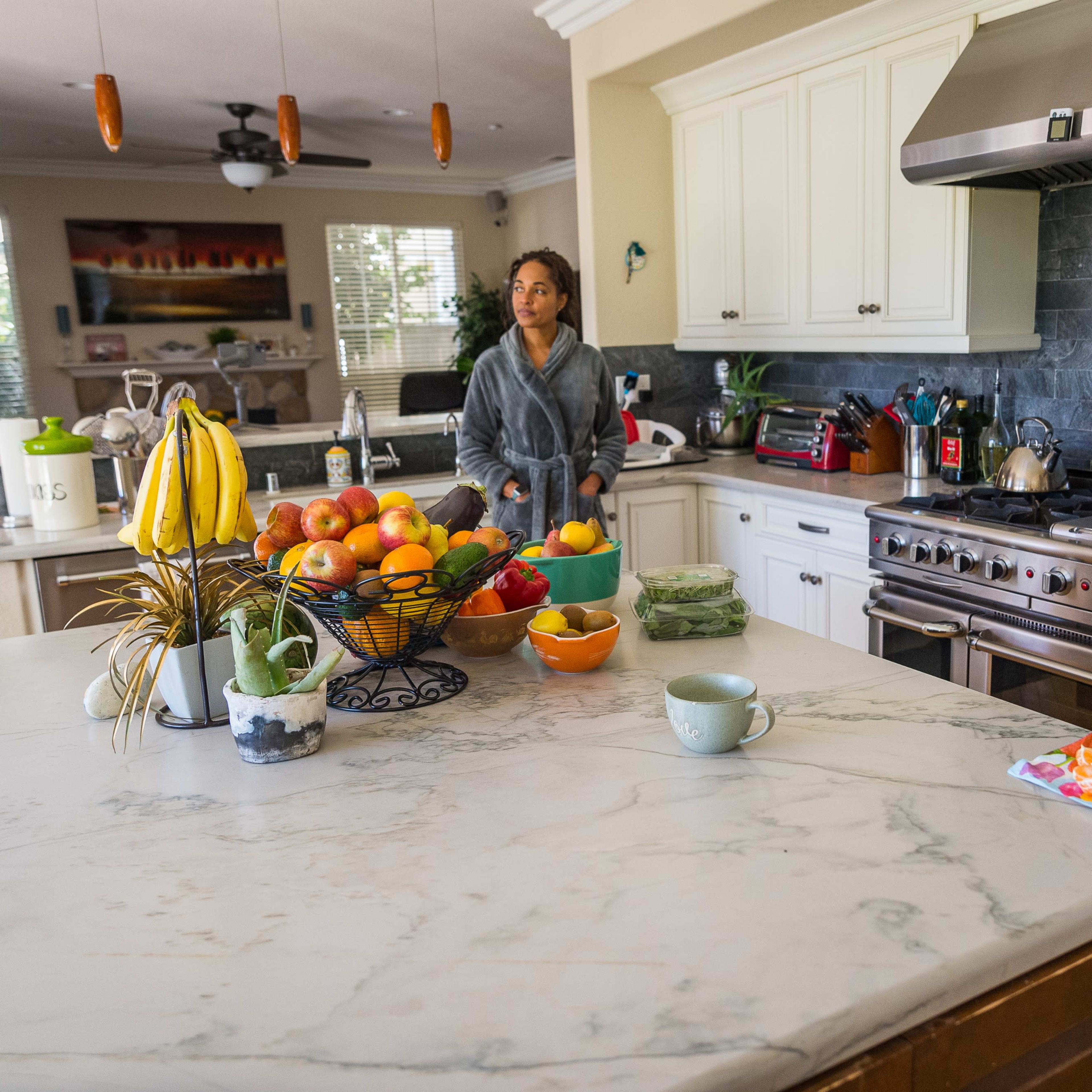 A person stands in a kitchen with a marble countertop, surrounded by bowls of fresh fruit and a bowl of vegetables, near a stove and various kitchen appliances.