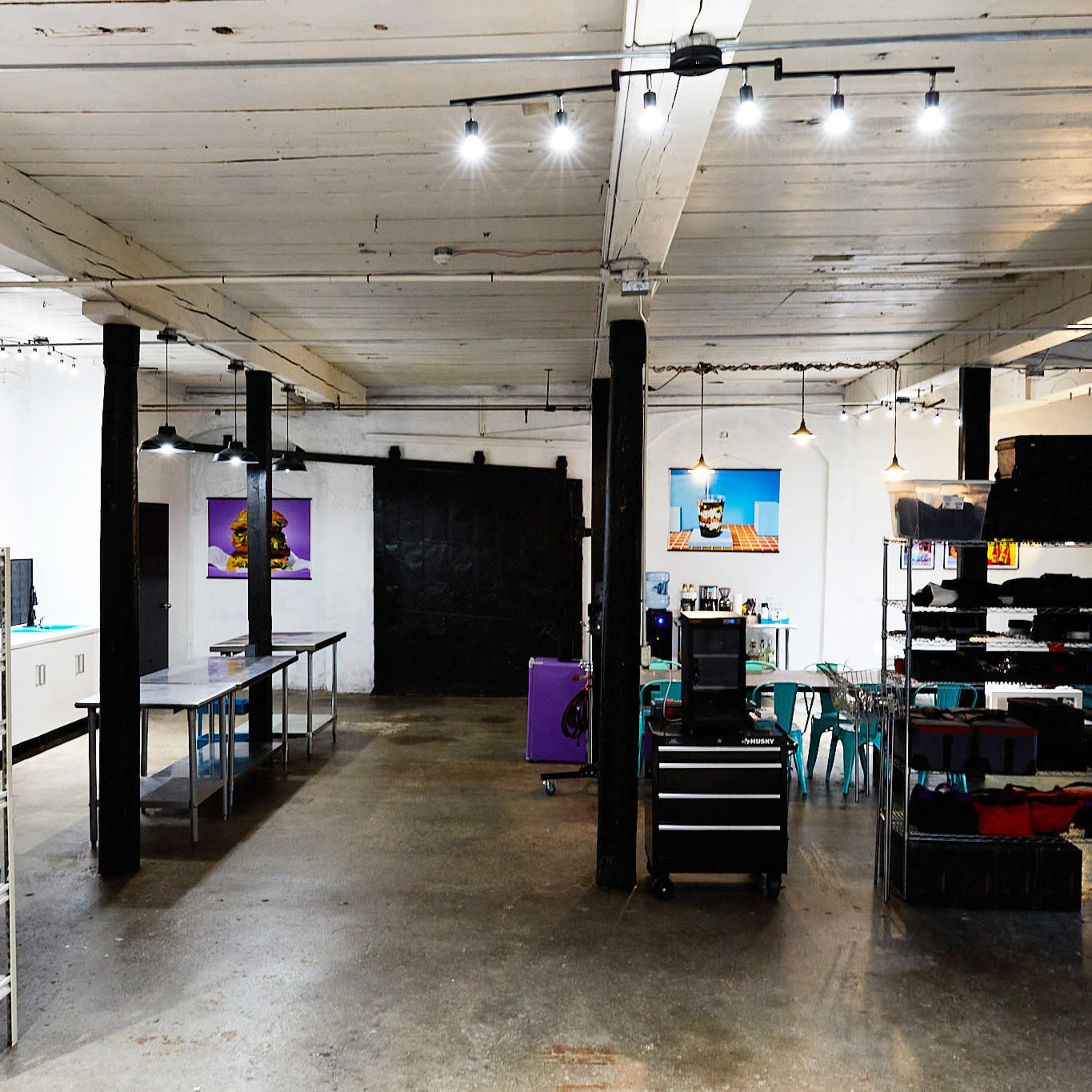 A spacious, industrial-style room features tables and shelves filled with various kitchen items, while art hangs on the walls and concrete flooring provides a minimalist backdrop.