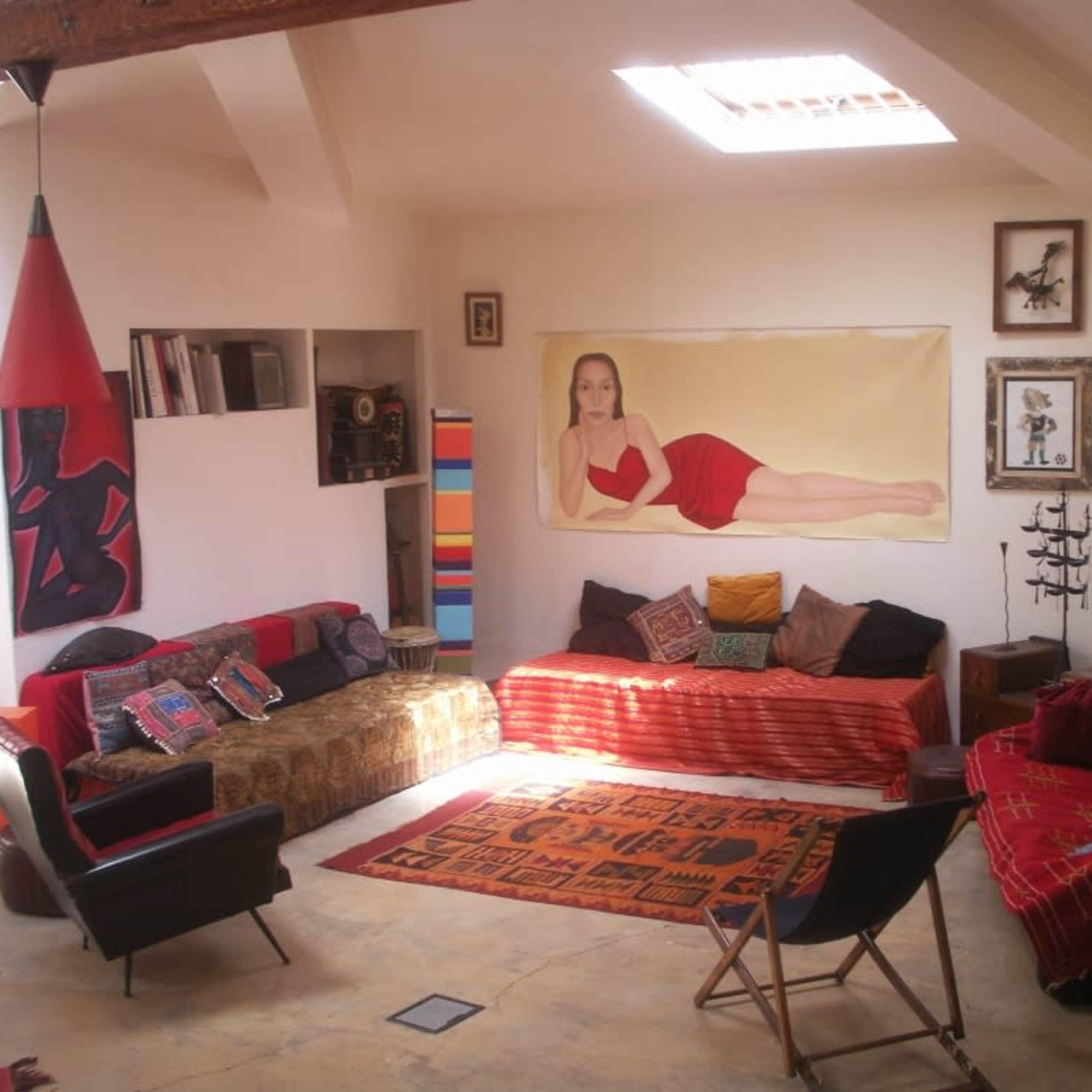 A sunlit living room featuring colorful couches, various wall art, and a mix of decorative pillows on the floor.