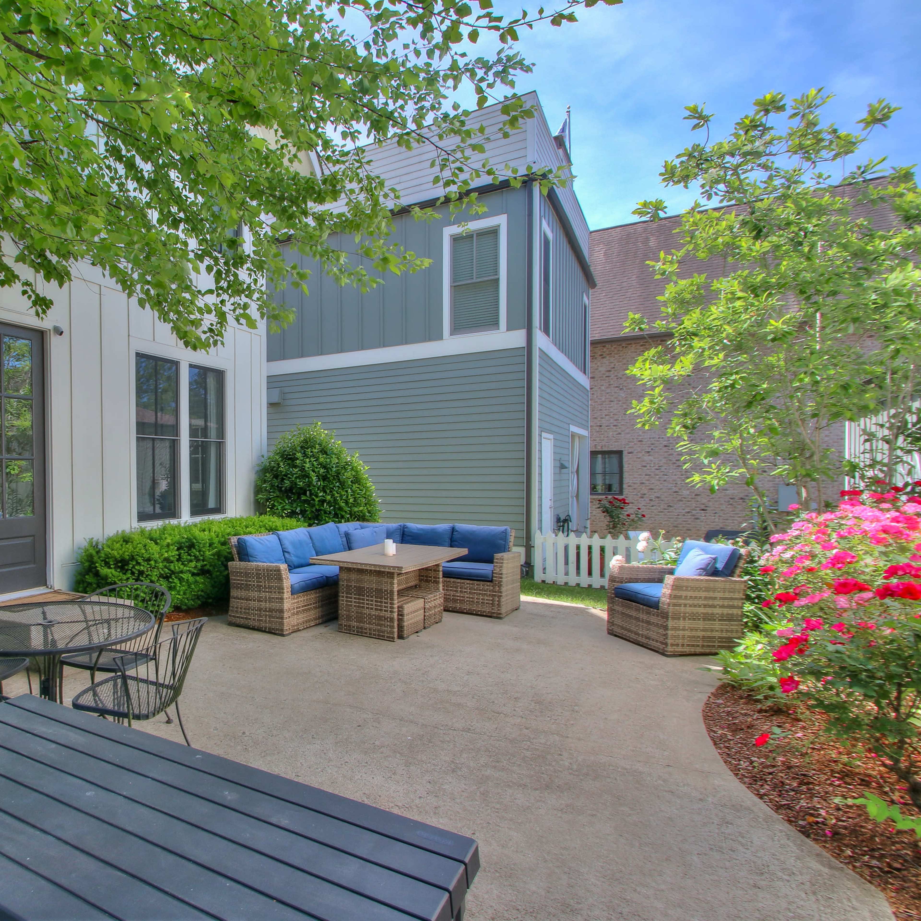 A patio features a seating arrangement with blue cushions and a small table surrounded by greenery and blooming flowers.