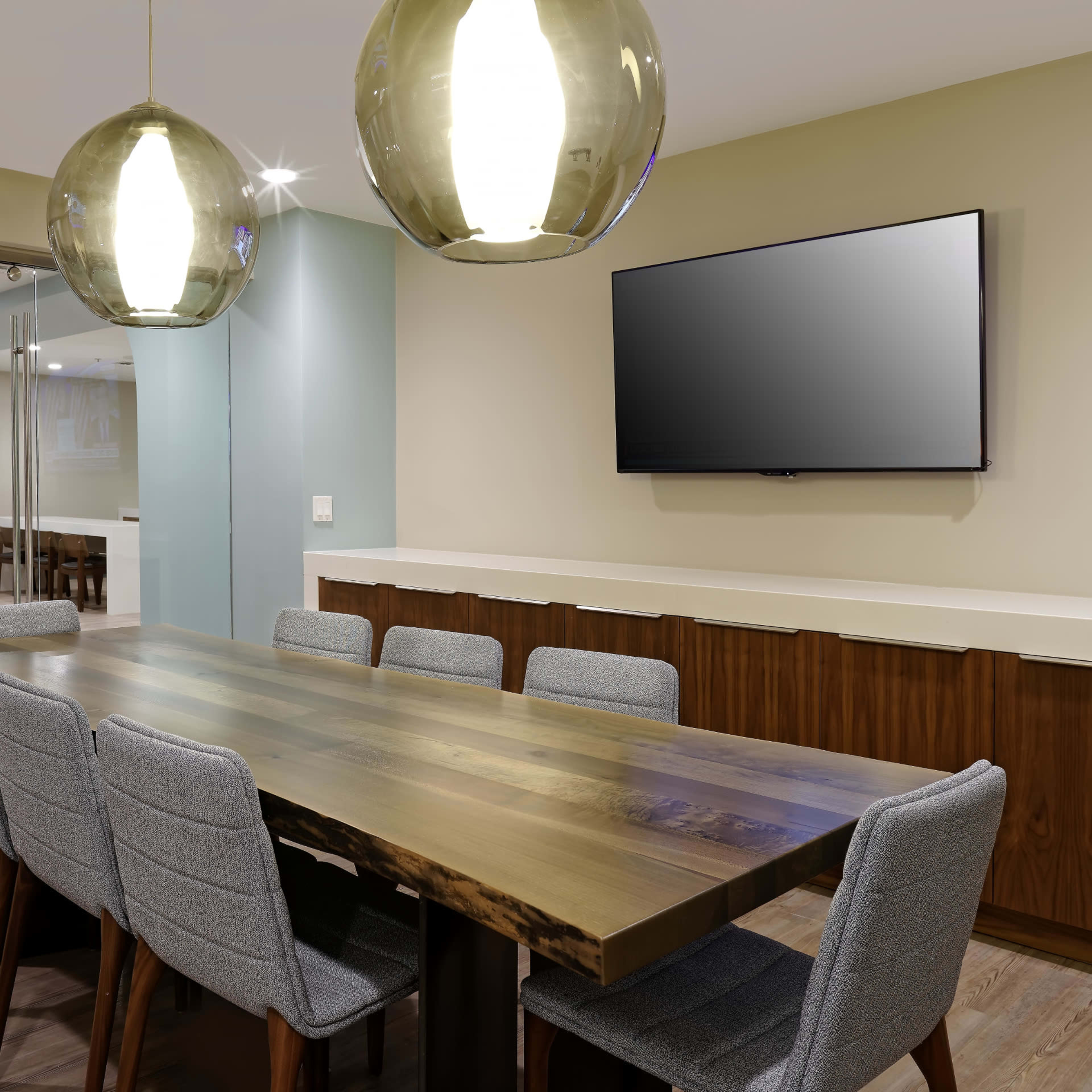A modern conference room features a long wooden table surrounded by gray upholstered chairs, with large pendant lights overhead and a wall-mounted television.