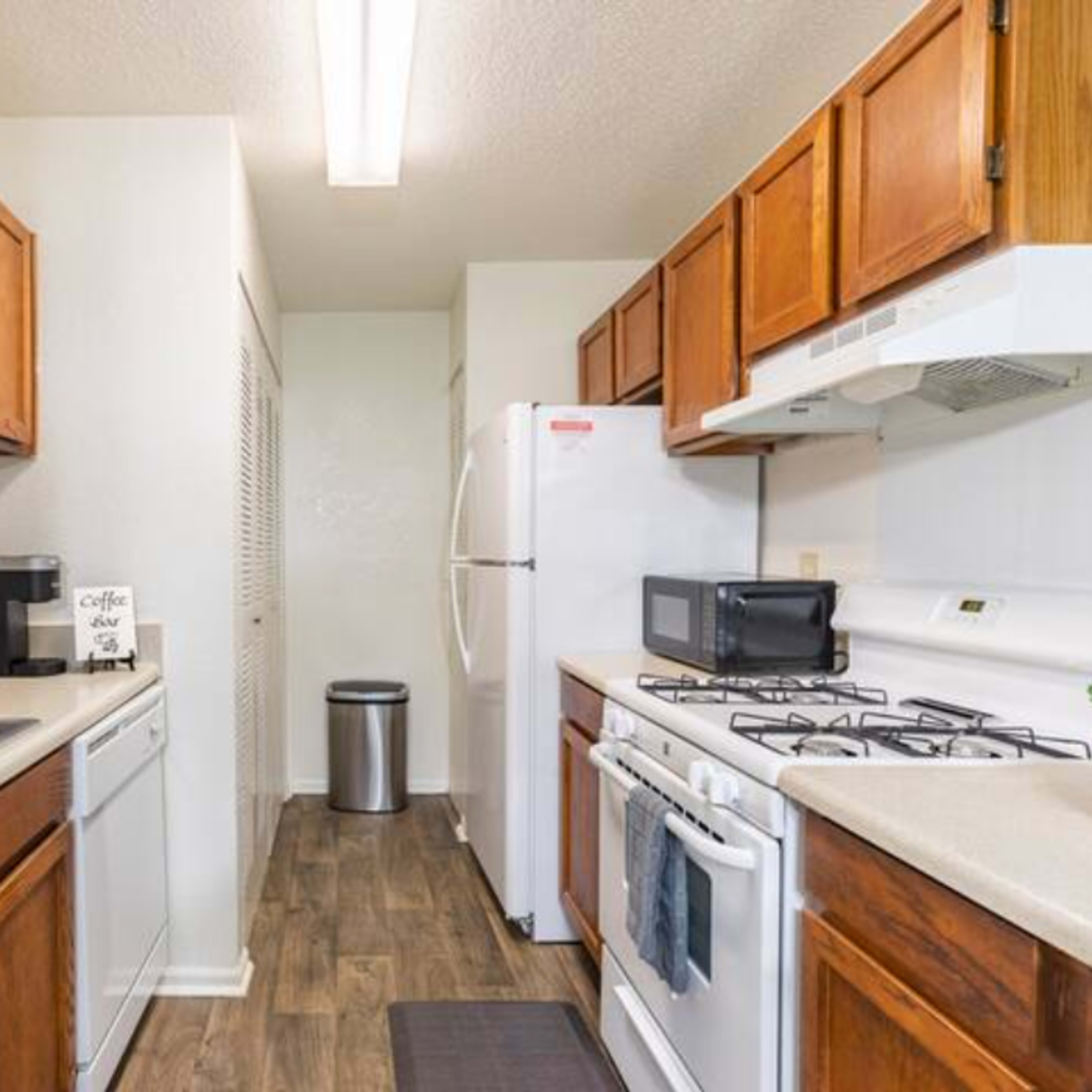 A narrow kitchen features wooden cabinets, a white refrigerator, a stove, and a double sink, with a coffee maker and a small microwave on the counter.