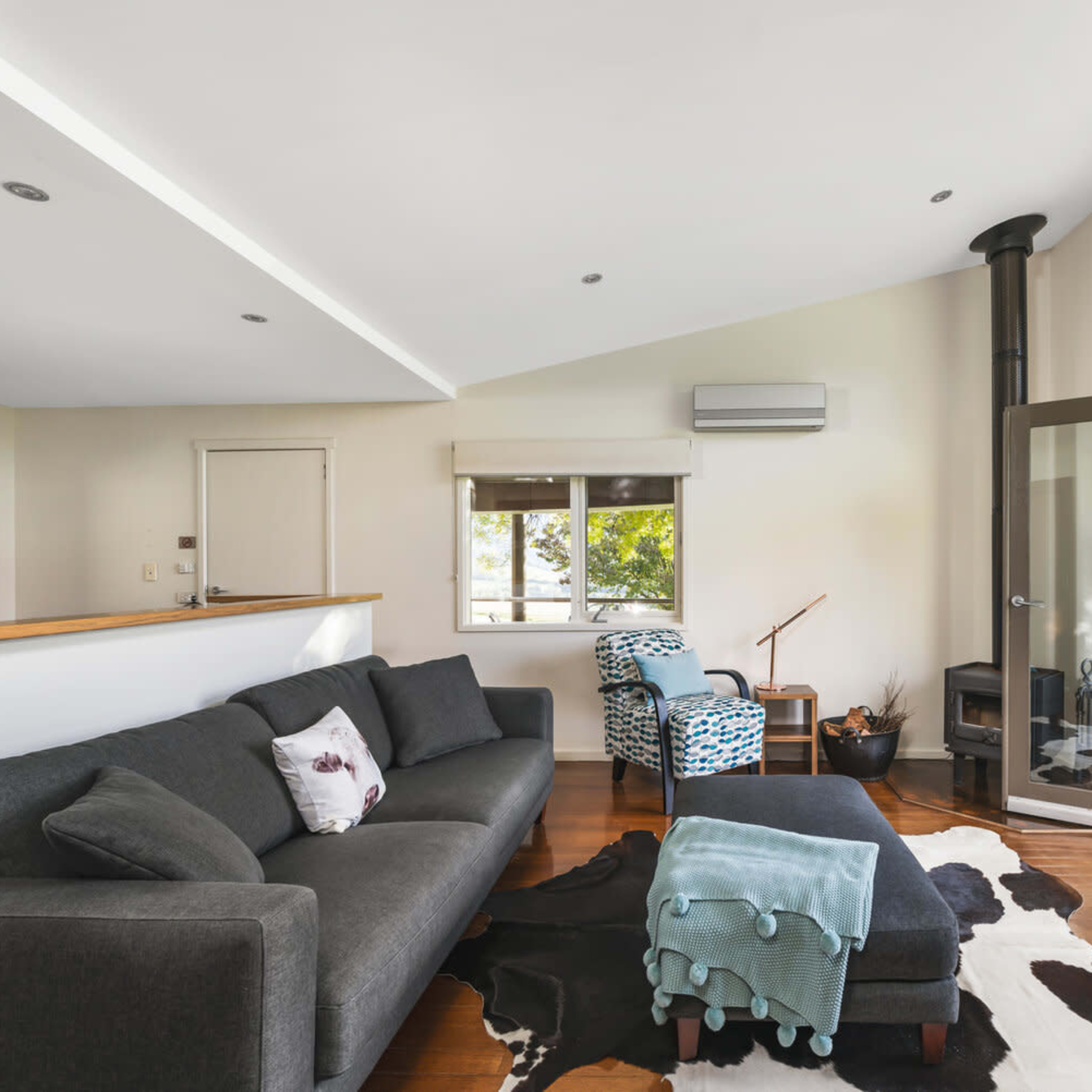 A modern living room with a dark sectional sofa, a patterned armchair, a wood-burning stove, and large windows opening to a deck.
