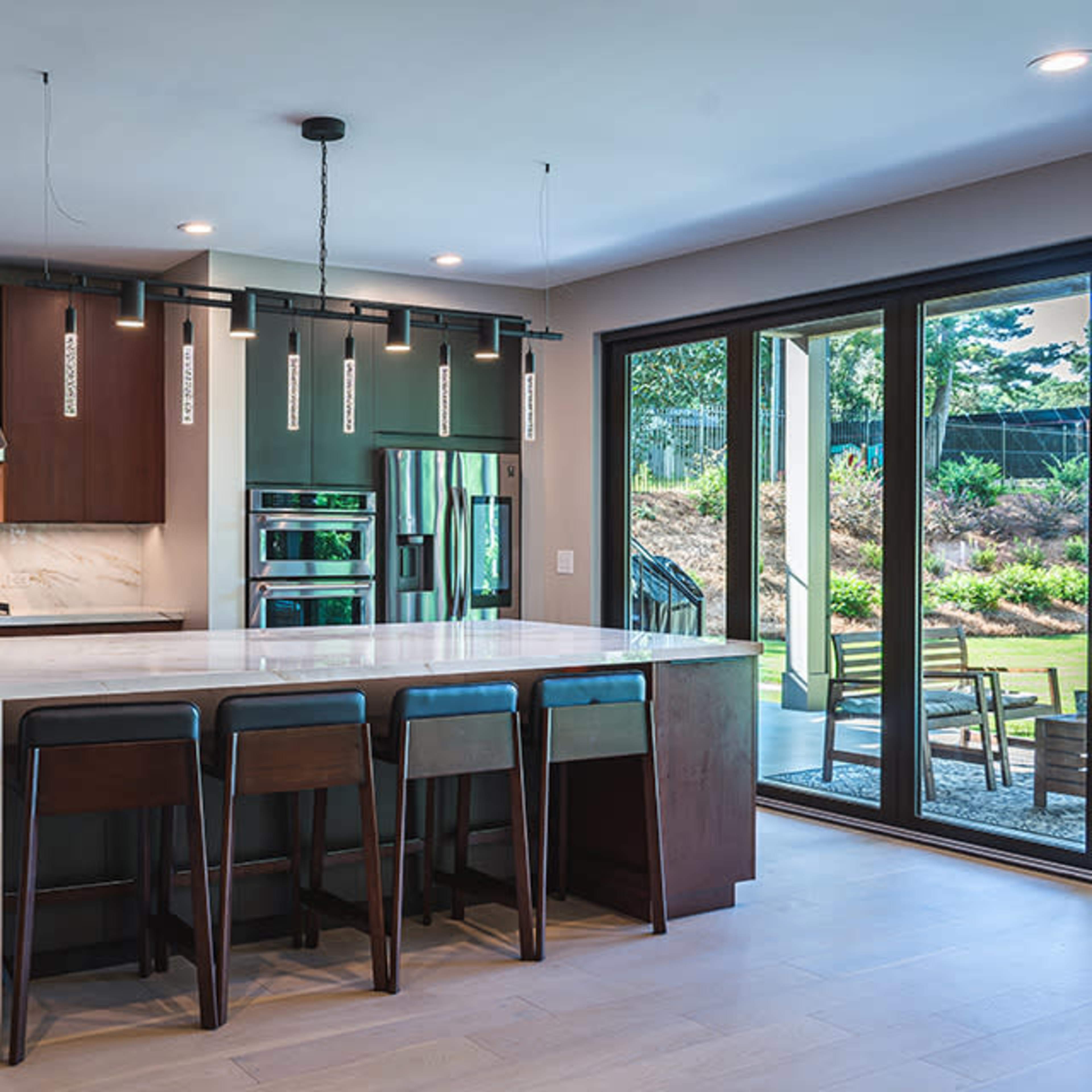 A modern kitchen features a large island with bar stools, dark cabinetry, and sliding glass doors leading to an outdoor patio.