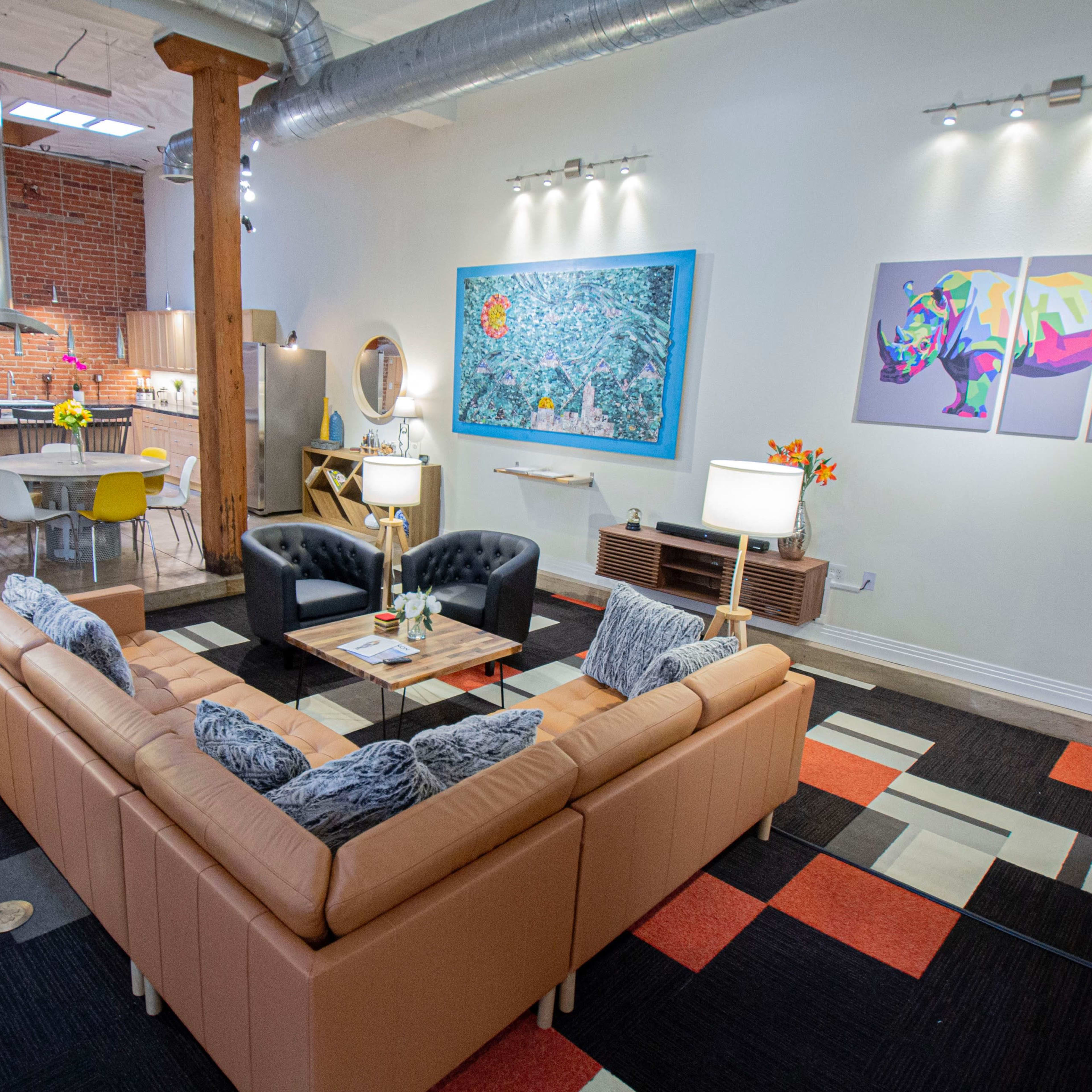 A modern living space featuring a tan sectional sofa, a colorful area rug, and artwork on the walls, with a view of a kitchen and dining area in the background.