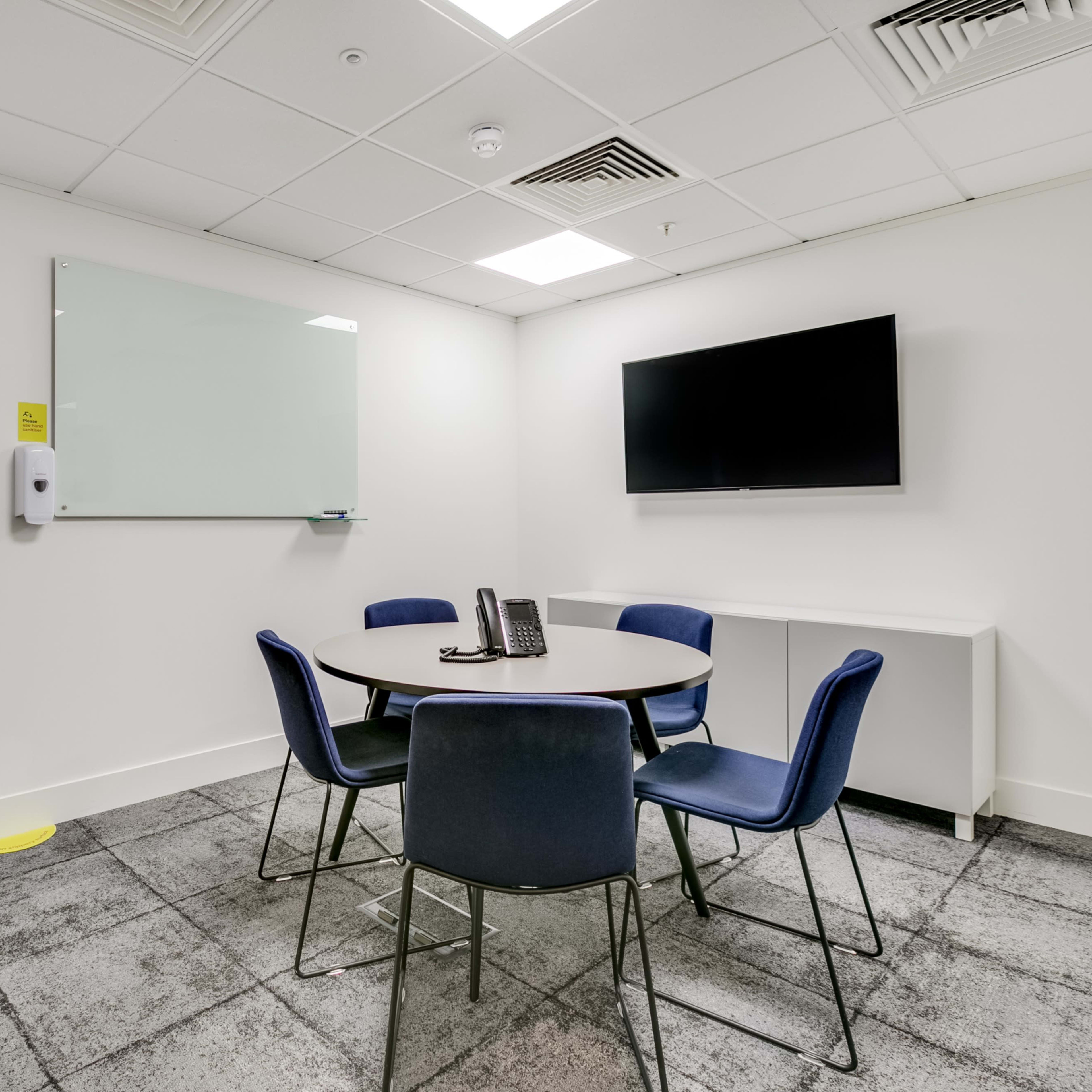A small meeting room features a round table with four chairs, a wall-mounted television, a whiteboard, and a plant in a corner.