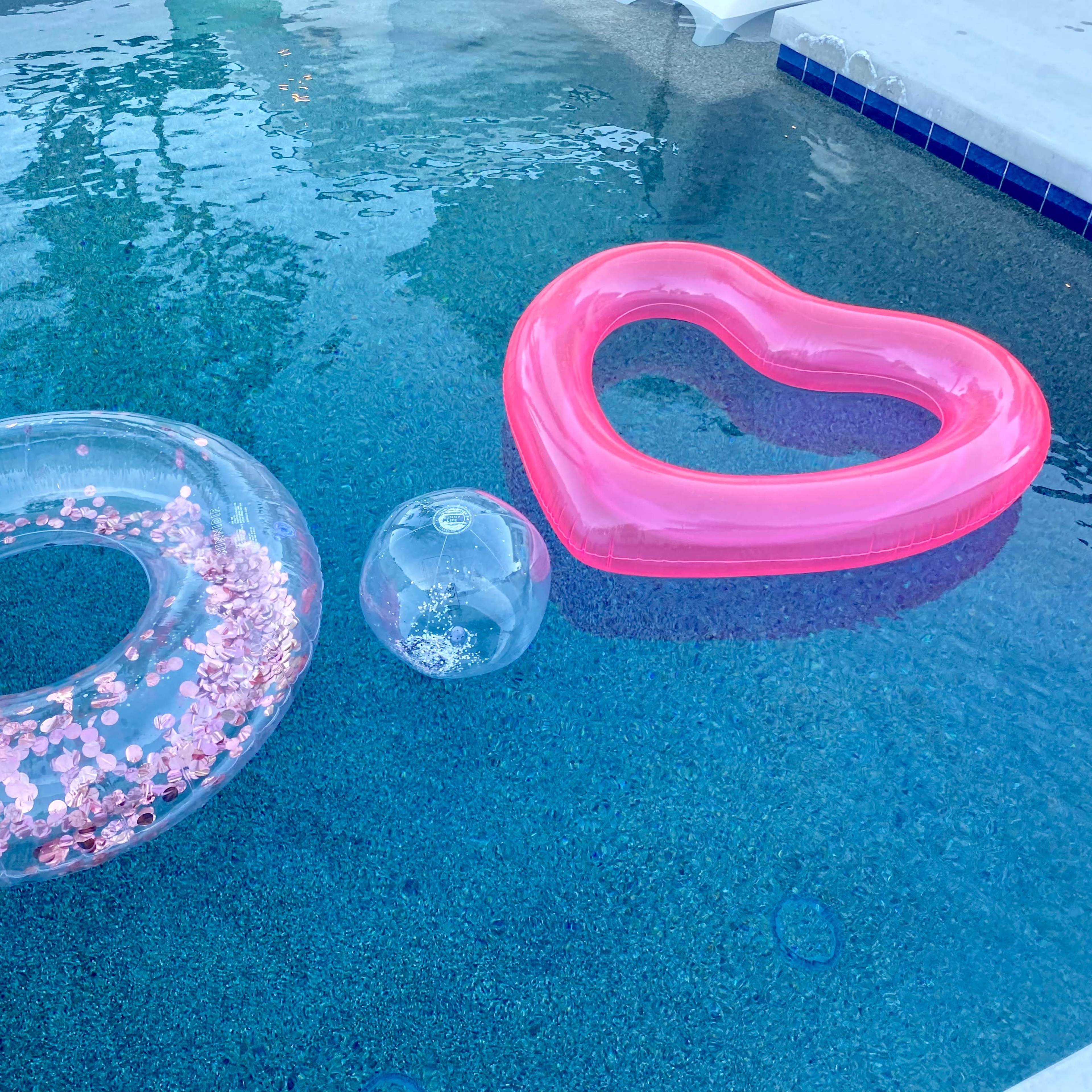 Three inflatable pool floats, including a transparent donut with pink confetti, a clear beach ball, and a pink heart-shaped float, are resting on the surface of a swimming pool.