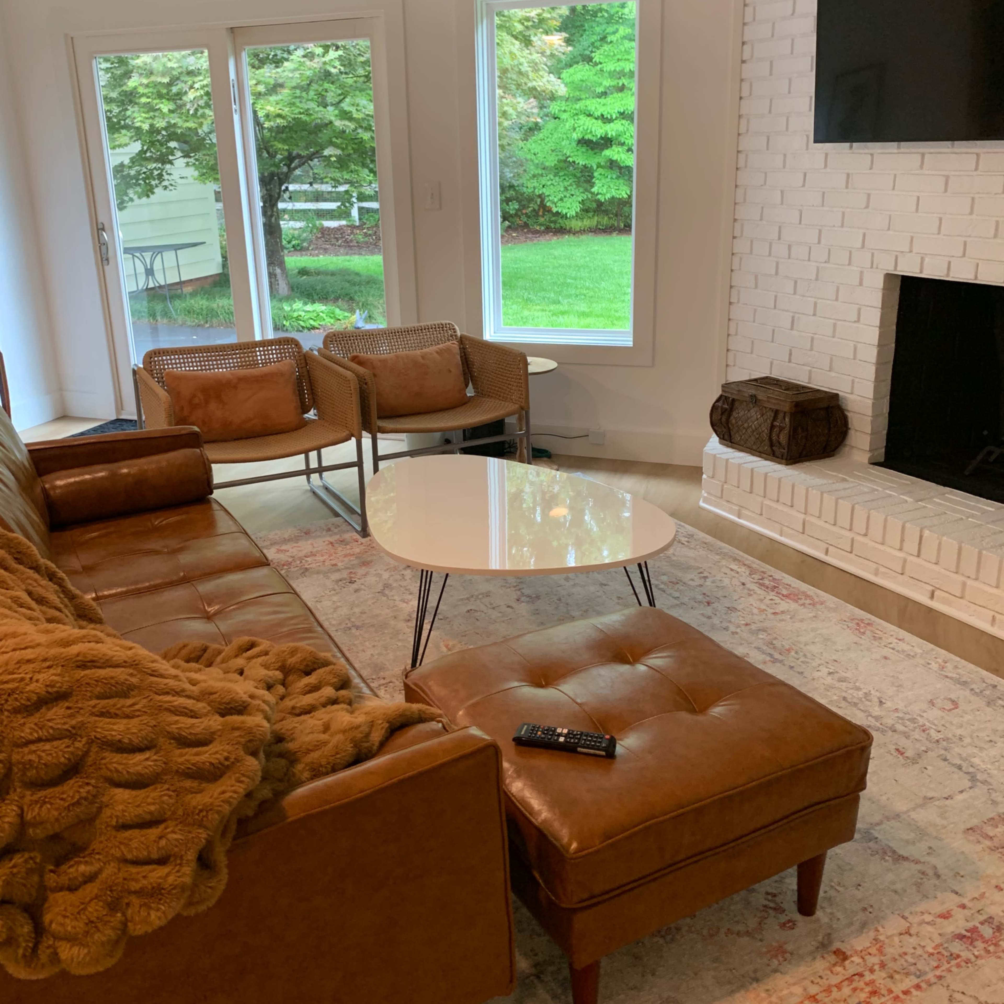 A modern living room features a brown leather sectional sofa, two woven chairs, and a round coffee table in front of a fireplace and large windows overlooking a green yard.