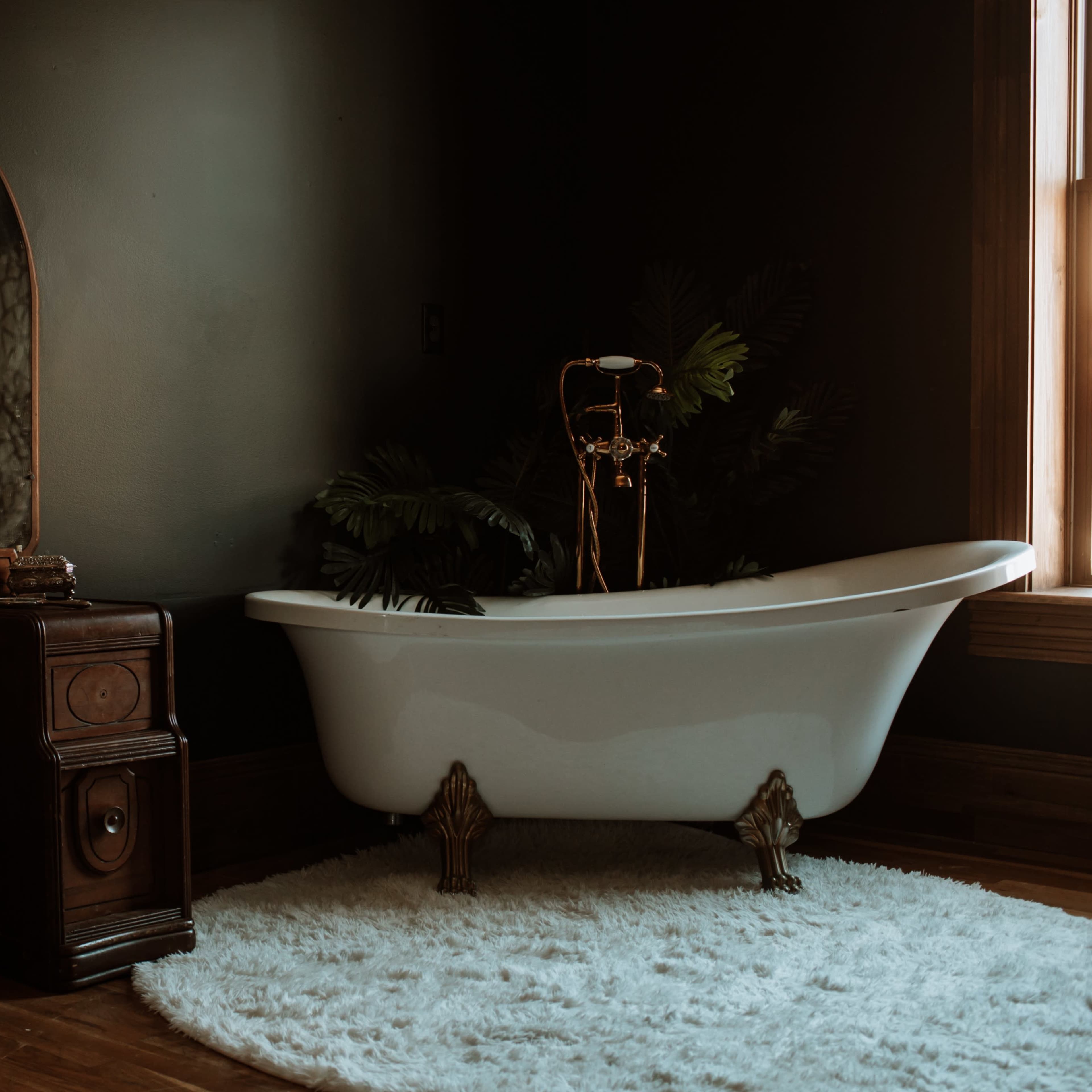 A vintage bathroom features a freestanding white bathtub beside a wooden vanity with a large mirror, all set against a dark wall.