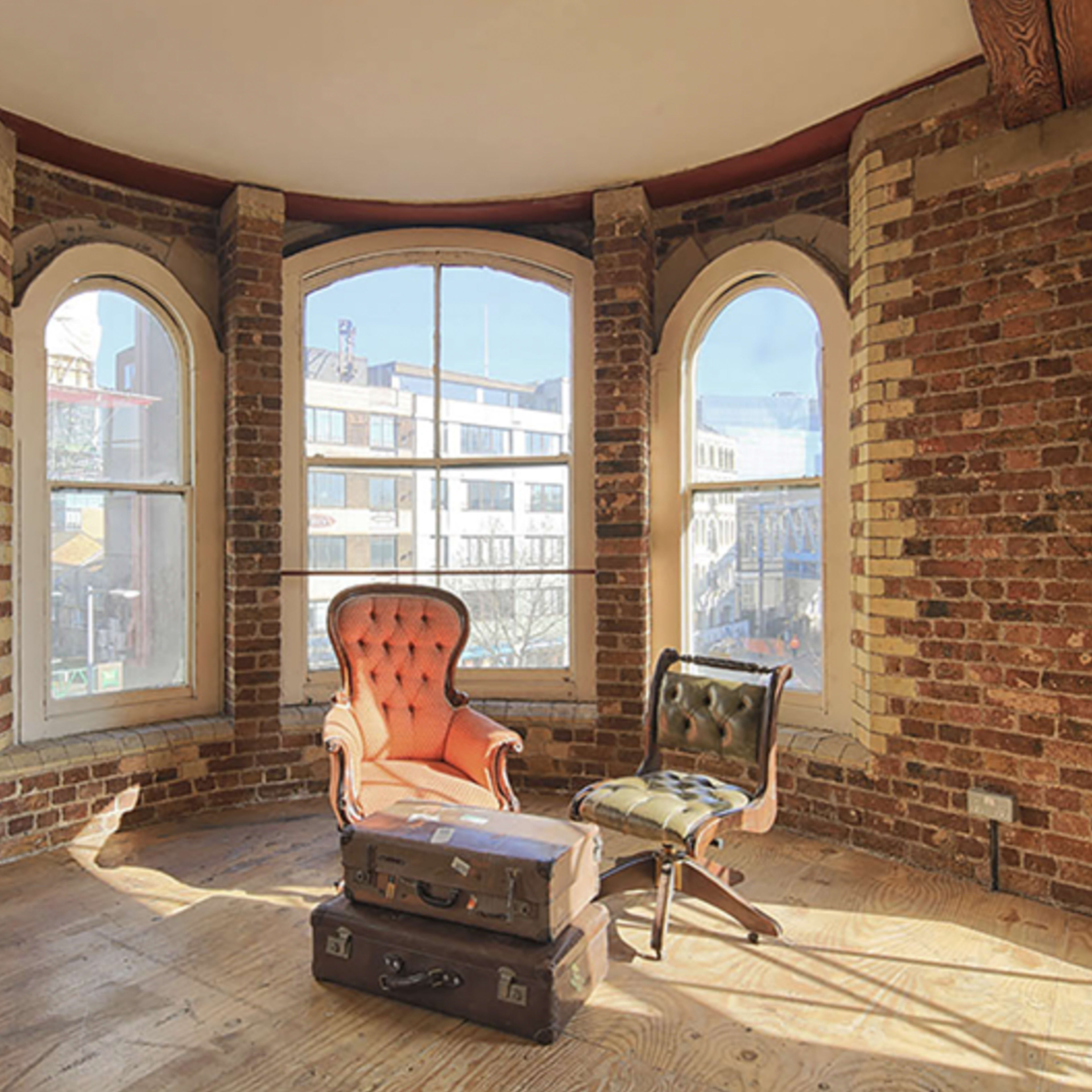 A cozy corner with a brick wall featuring two armchairs and two vintage suitcases on a wooden floor, illuminated by natural light through large windows.