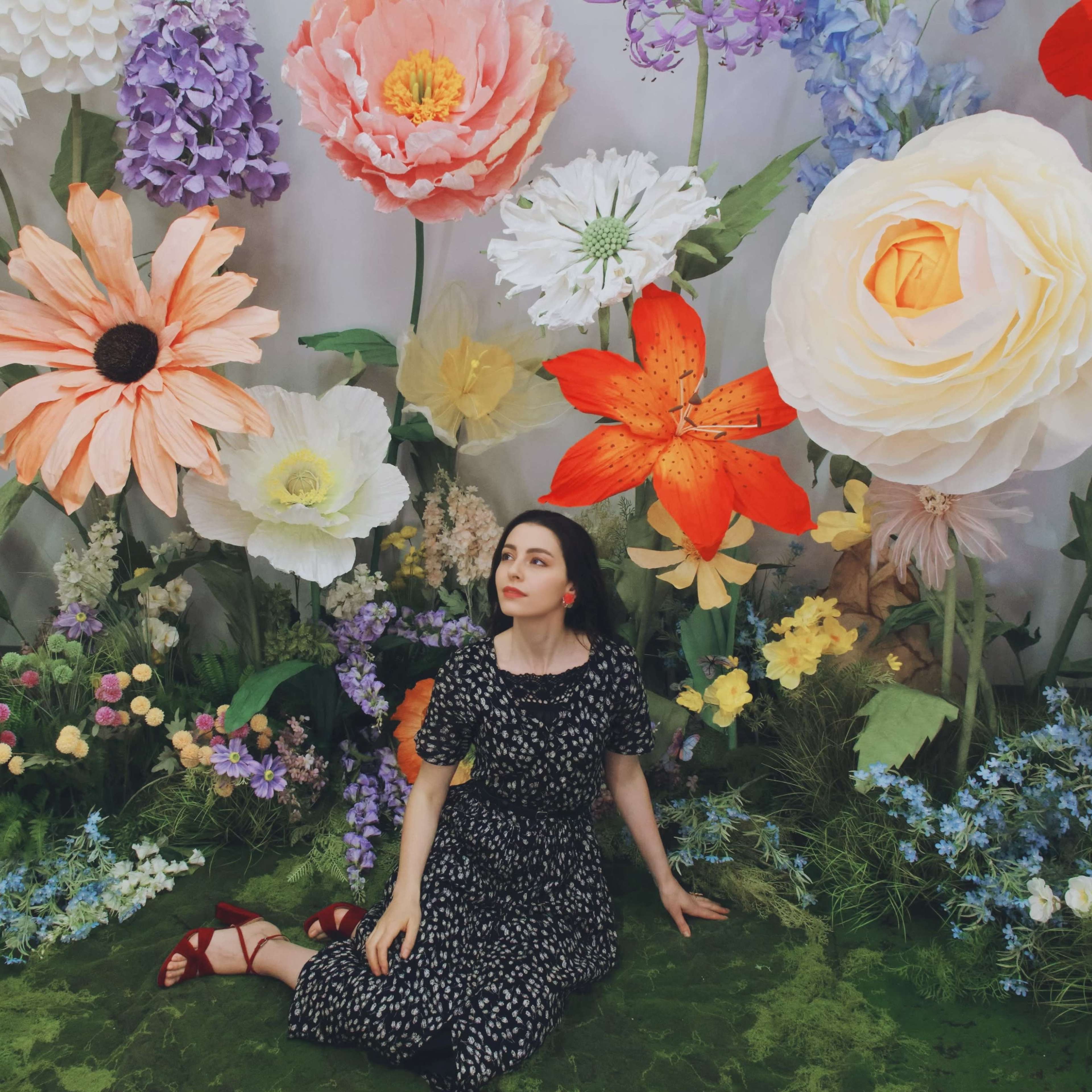 A woman sits among large, colorful floral decorations in a vibrant garden setting.