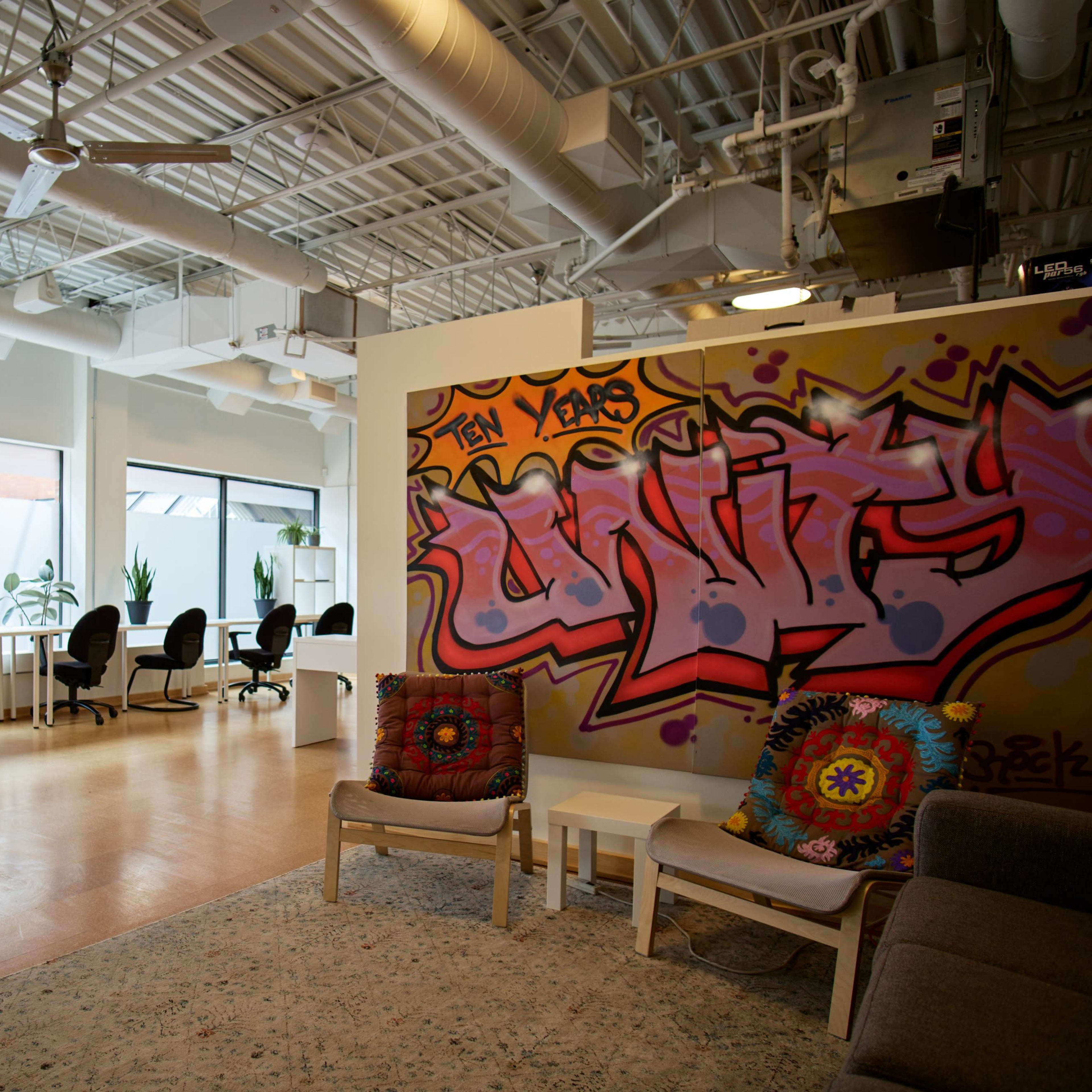 A modern workspace with a colorful graffiti wall, an area for seating, and several desks arranged in a well-lit room with plants and large windows.