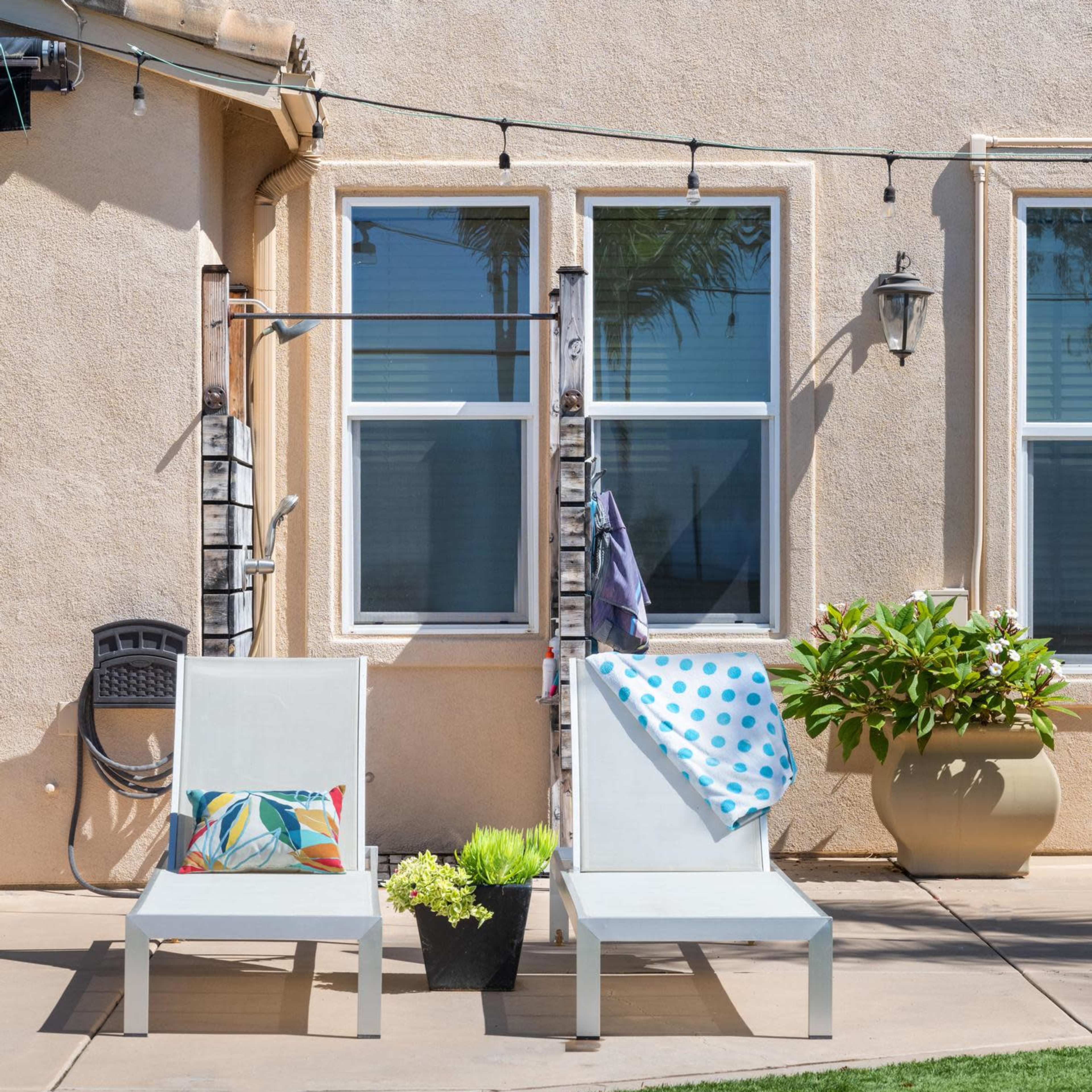 Two white lounge chairs are positioned by a poolside, with one chair featuring a colorful pillow and the other draped with a blue polka-dot towel, next to a potted plant.