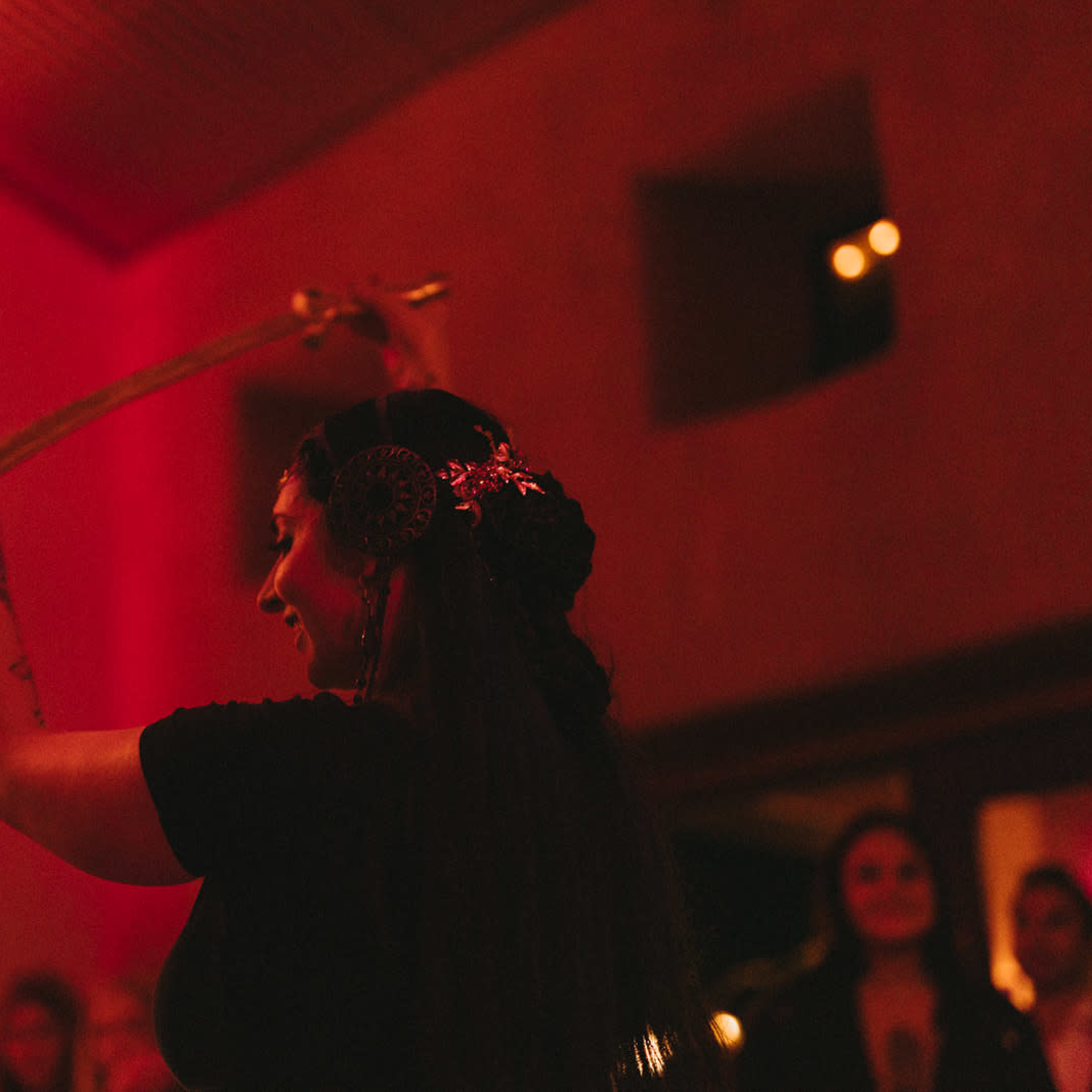 A woman in a black dress holds a sword above her head while performing in a dimly lit room with an audience visible in the background.