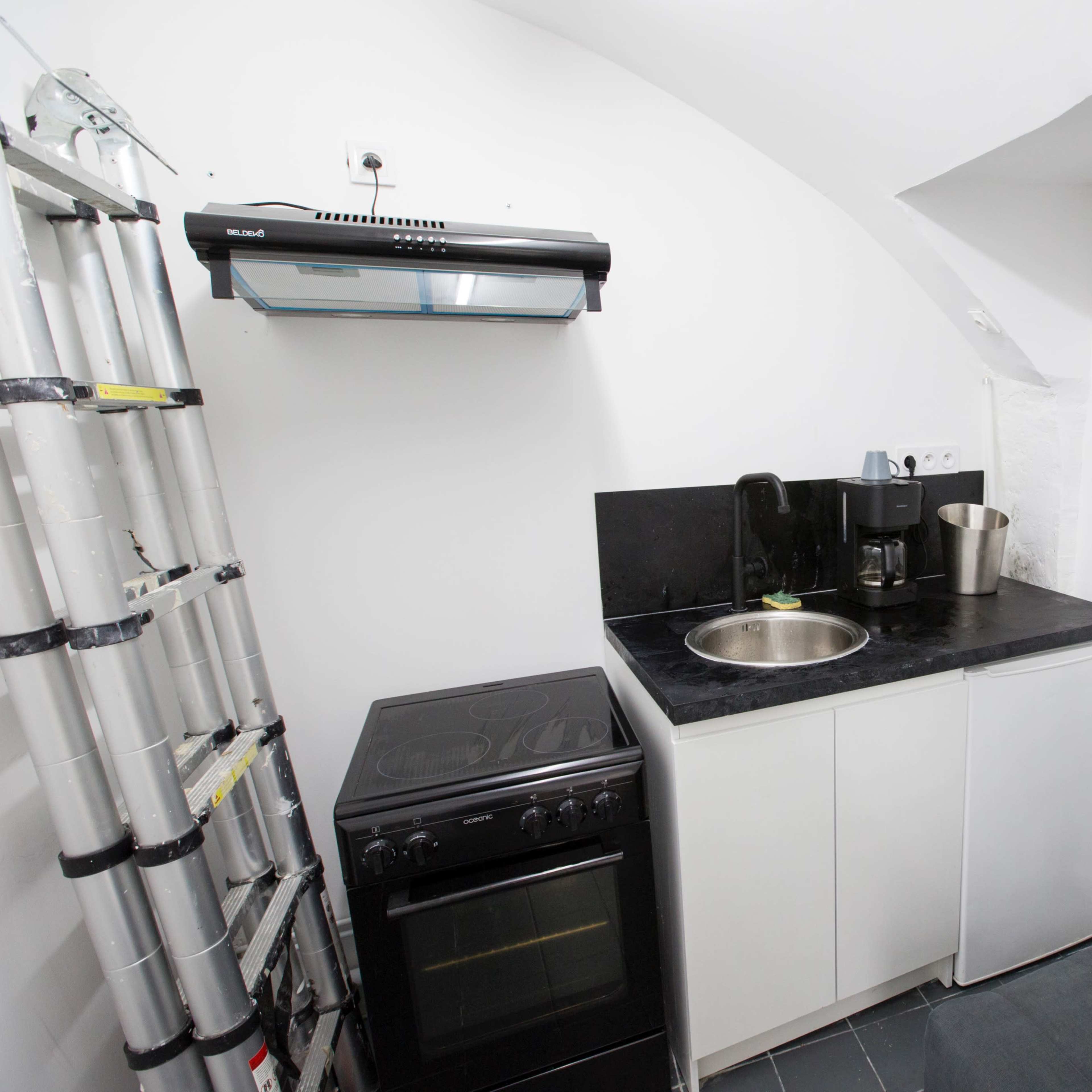 A small kitchen area featuring a sink, coffee machine, stove, and a gray couch against a white wall.