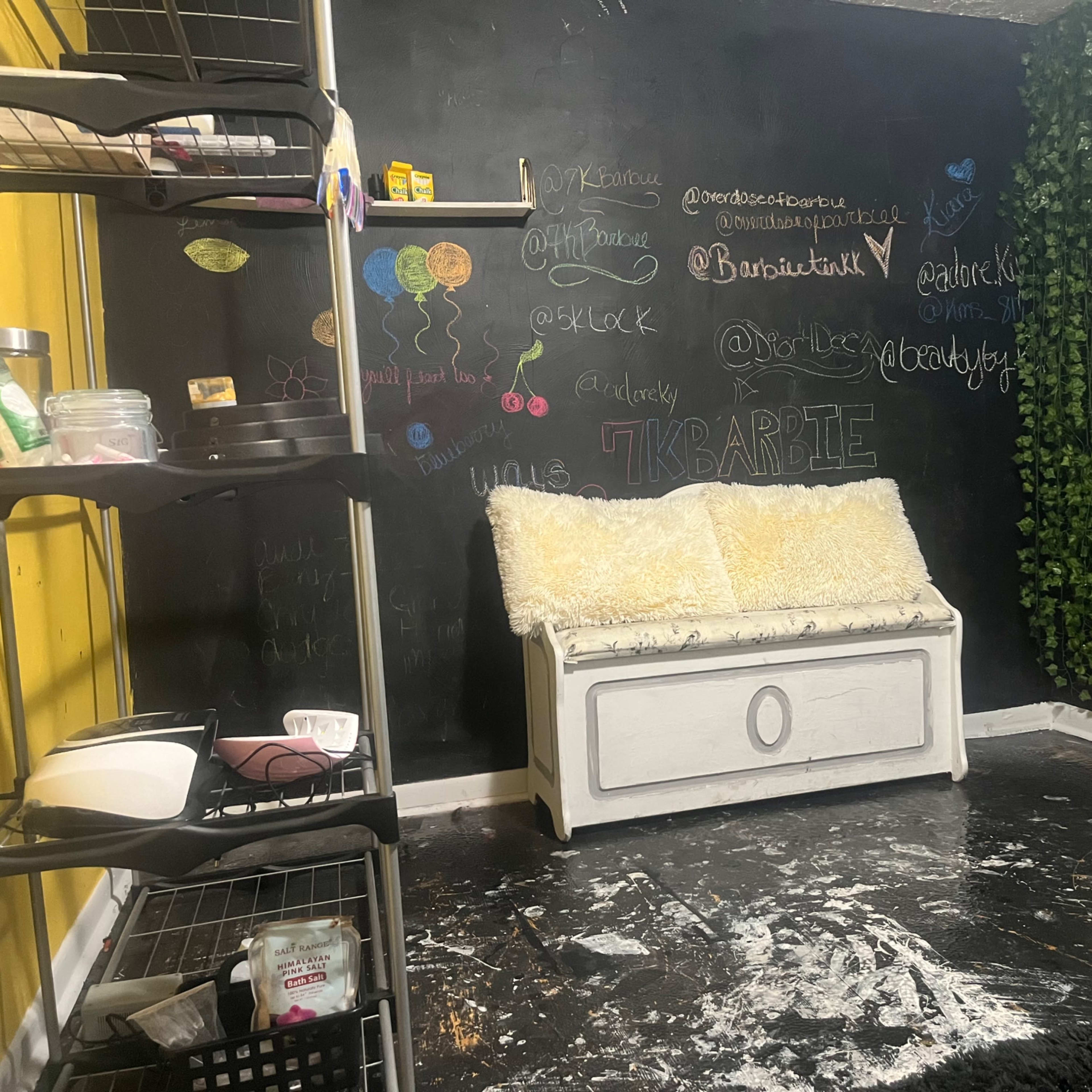 A room featuring a black chalkboard wall with various colorful writings, a white storage bench with a fluffy cover, and a section of greenery alongside a black door.