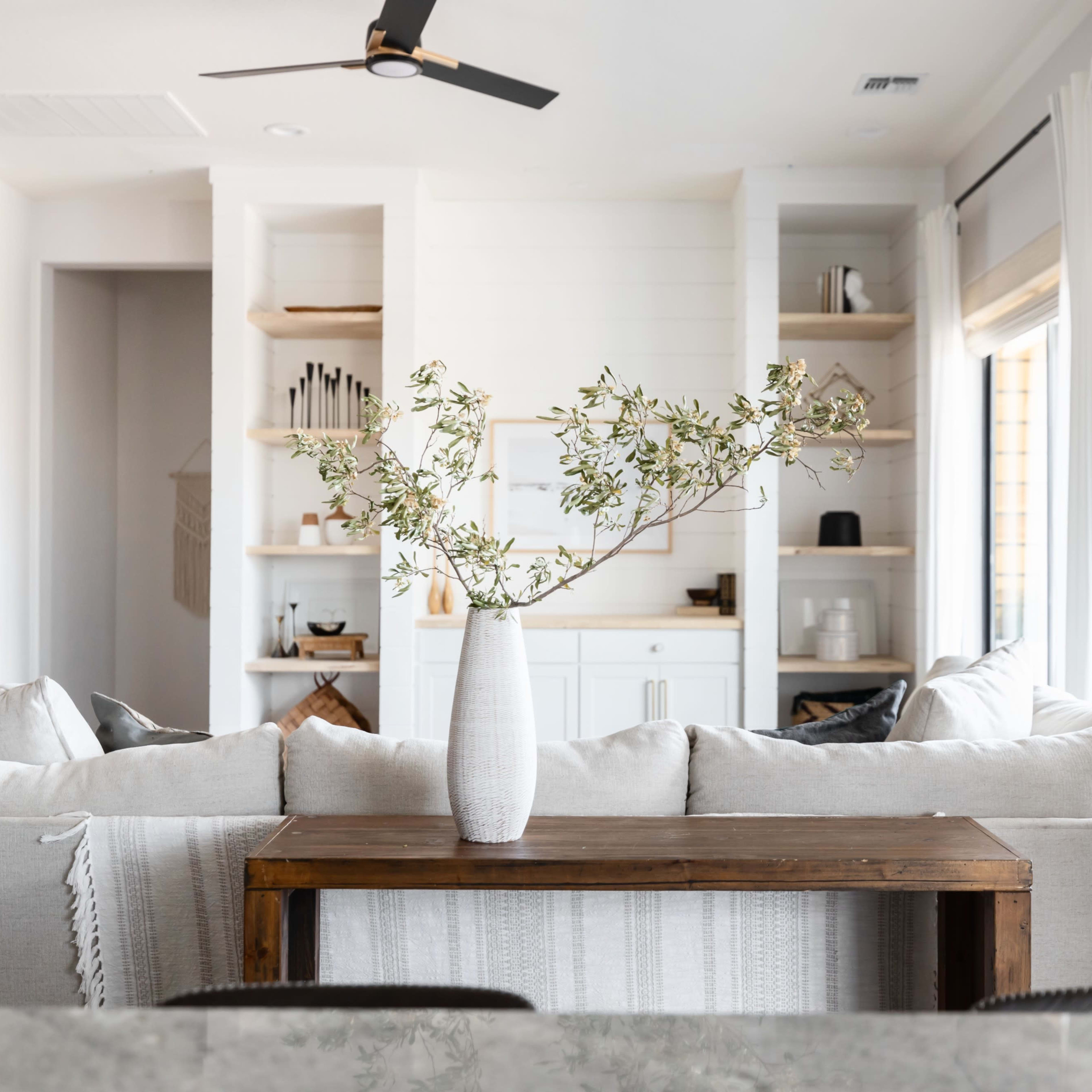 A spacious living area features a neutral-colored sofa with a wooden table and a vase of greenery, overlooking built-in shelves and large windows.
