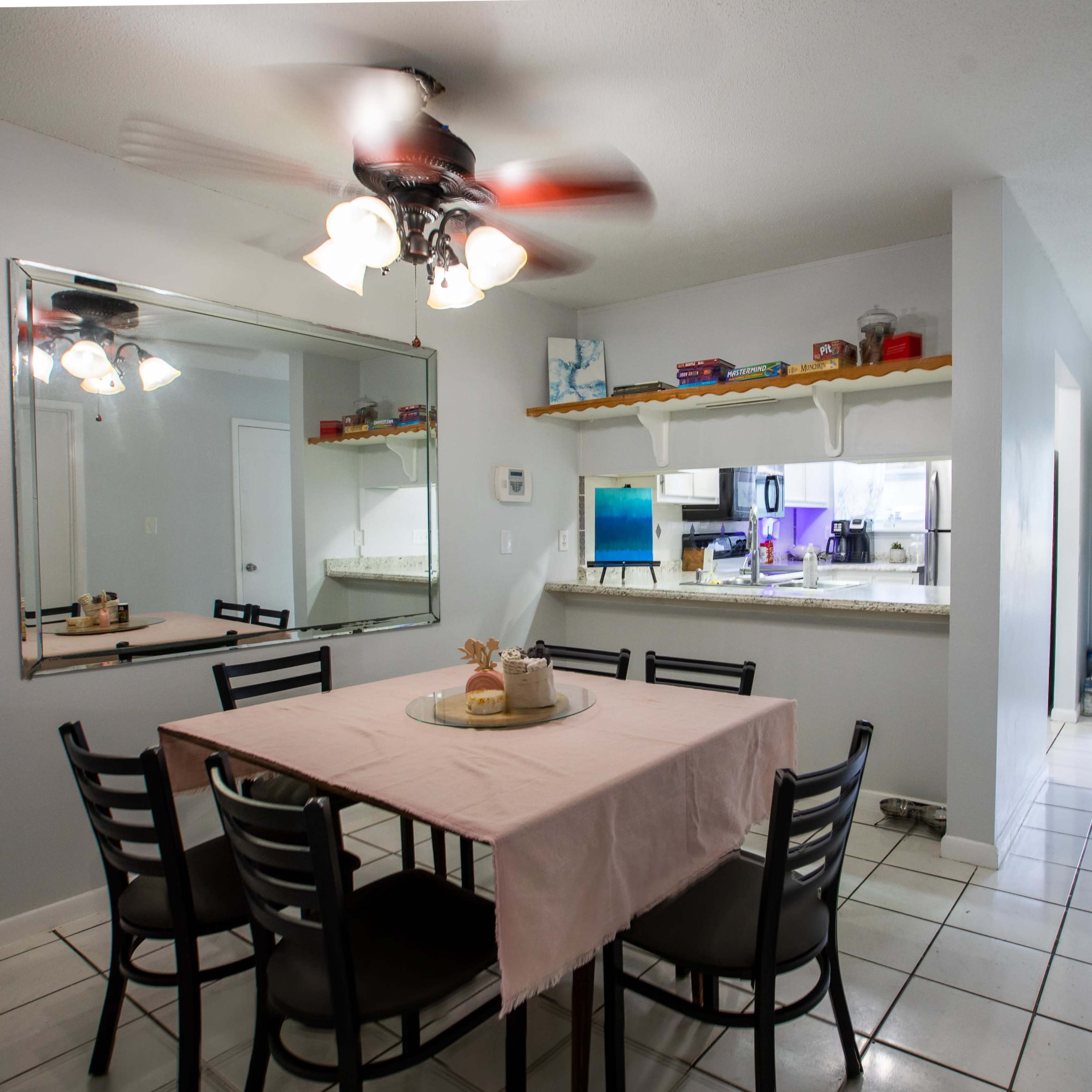 A dining area features a rectangular table with a pink tablecloth surrounded by black chairs, with a mirror and a view of a kitchen in the background.