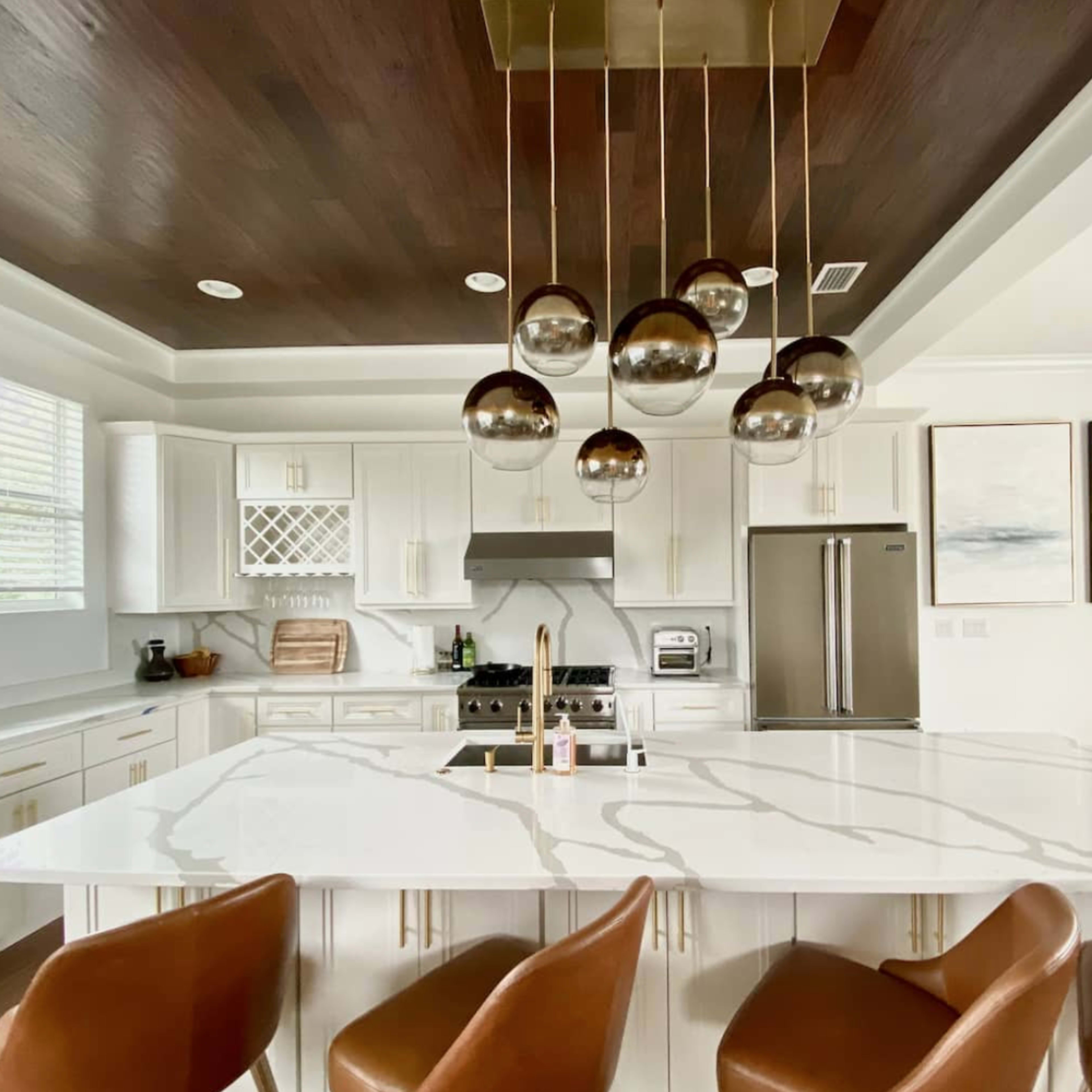 A modern kitchen features a large marble island with metal bar stools, pendant lighting, and stainless steel appliances.