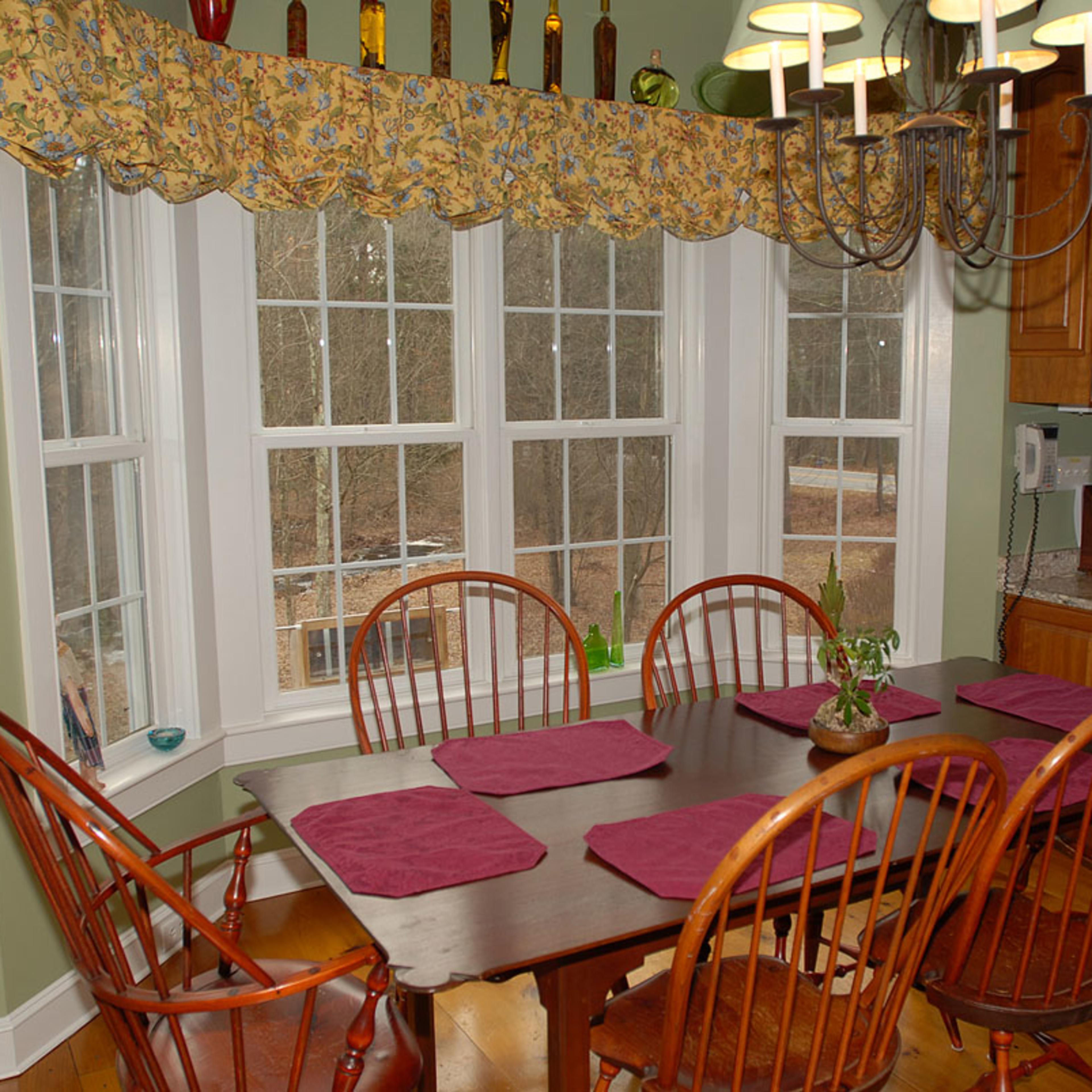 A wooden dining table with eight chairs is set in a well-lit kitchen area featuring large windows and wooden cabinetry.