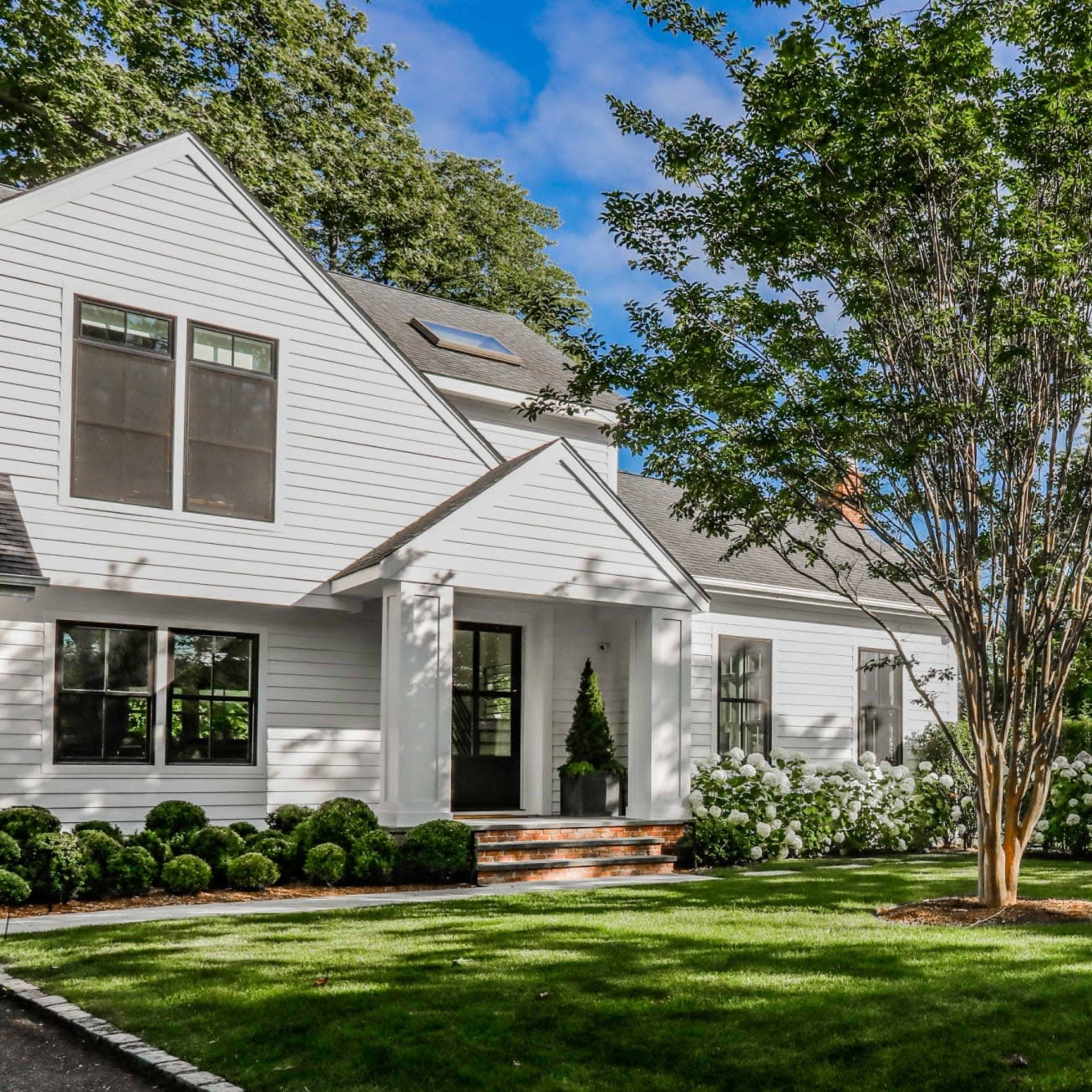 A modern white house with a landscaped front yard features a central tree and flowers lining the pathway.