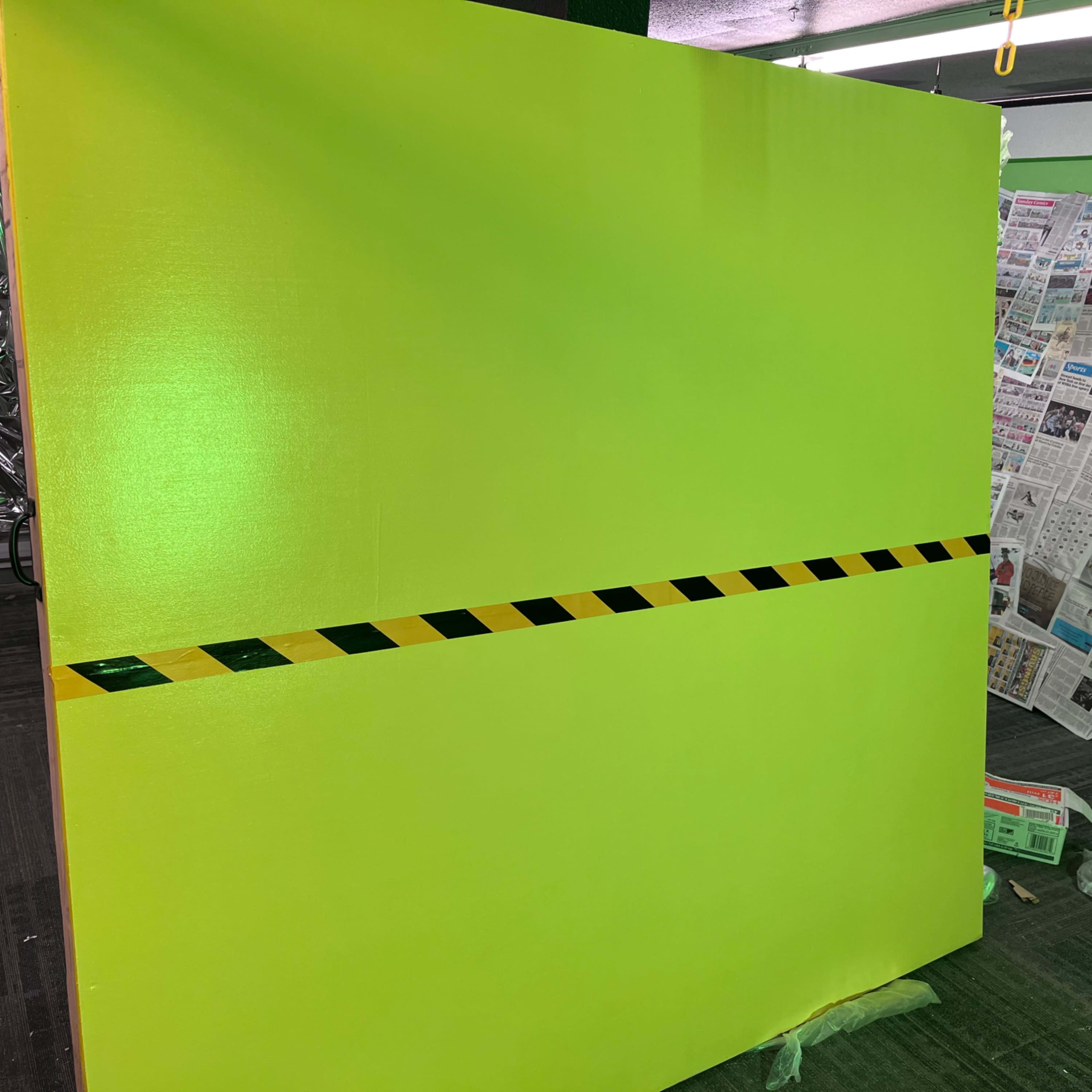 A large green wall with a black and yellow striped caution tape running horizontally across it stands in an indoor space, adjacent to a backdrop of newspaper clippings.