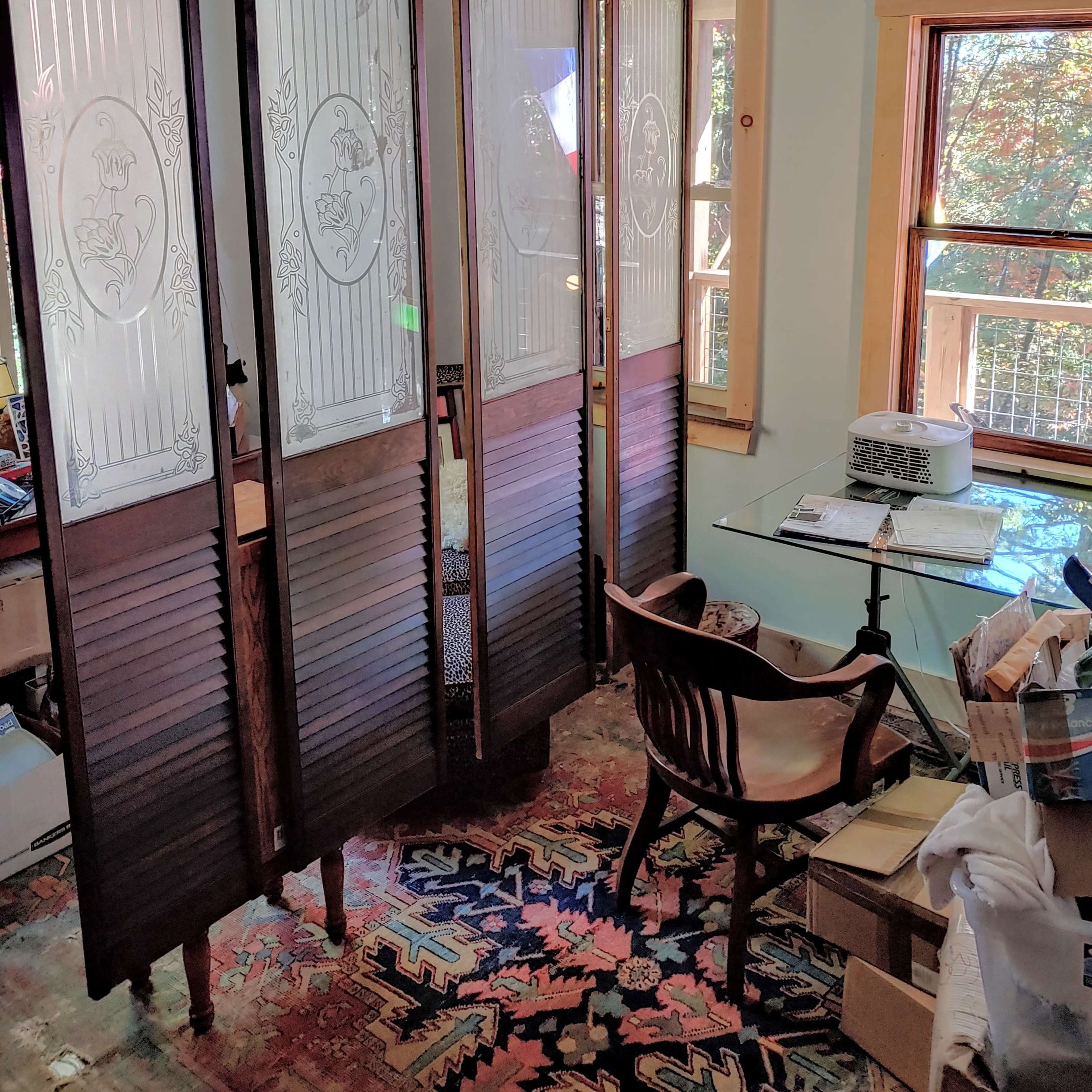 A wooden folding screen creates a divided space in a room with a desk and chair, surrounded by various items and a patterned rug.