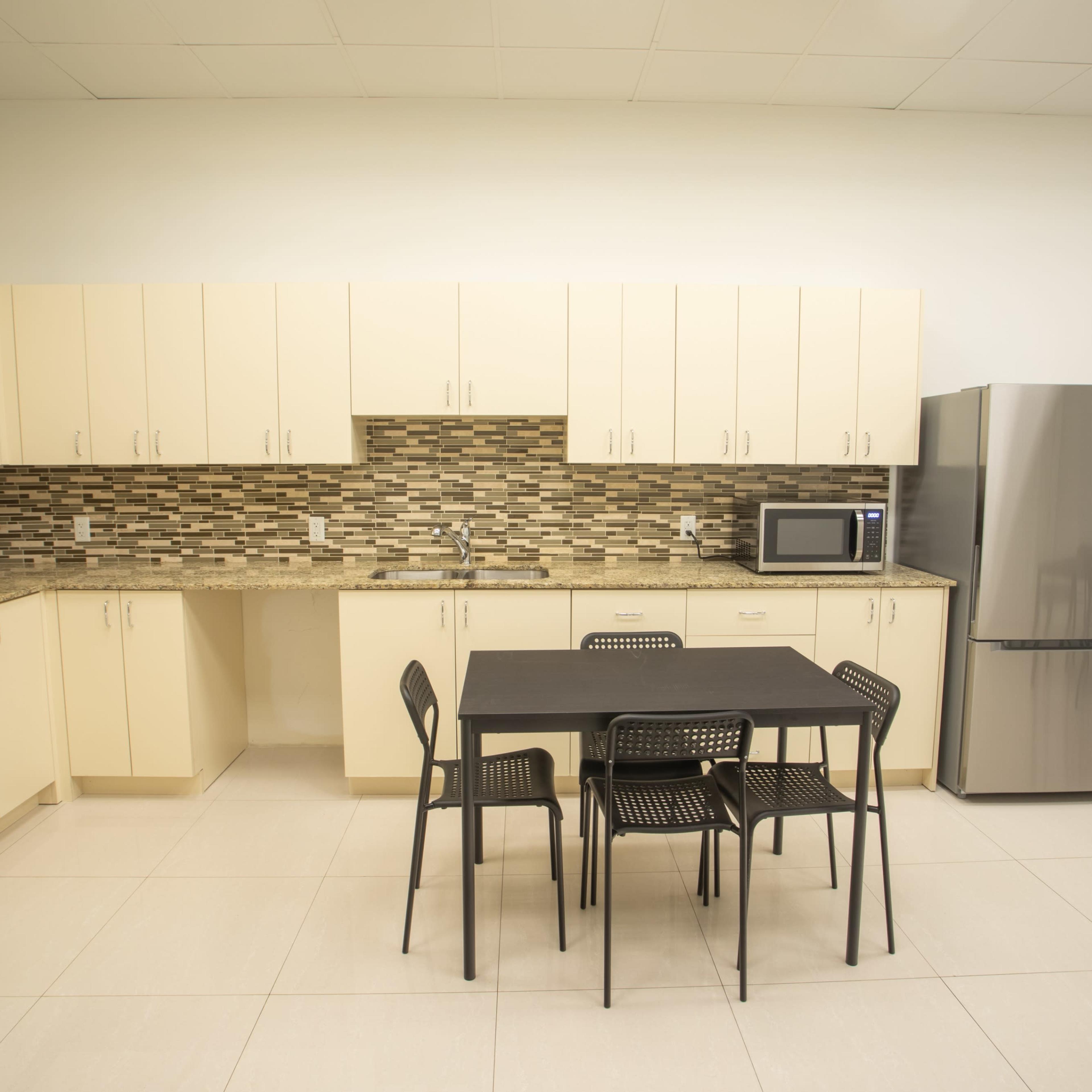 A modern kitchen features beige cabinets, a stone backsplash, a gray refrigerator, a microwave, and a small table with four black chairs.