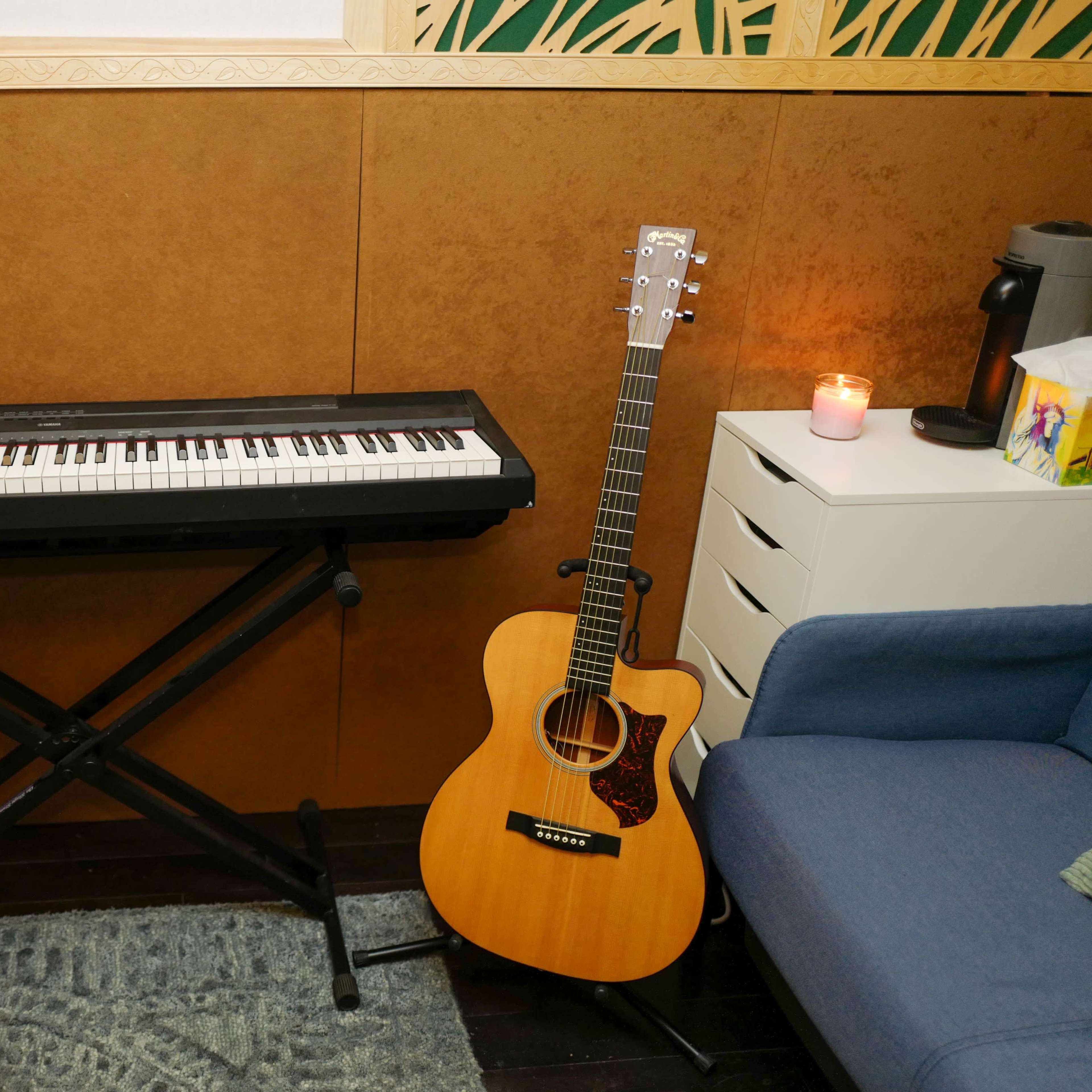 A digital piano on a stand next to an acoustic guitar and a lit candle in a room.