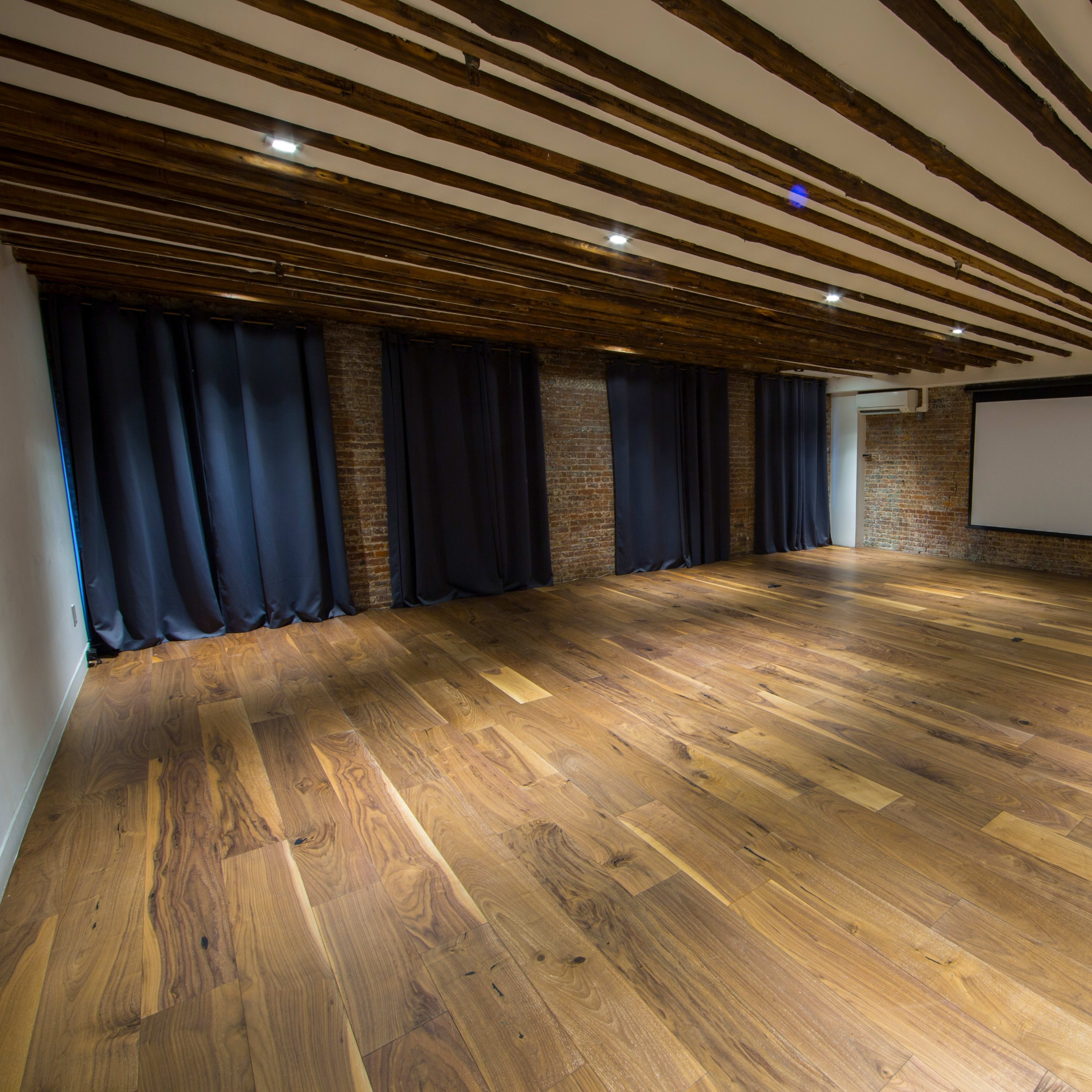 A spacious interior room with wooden flooring, exposed brick walls, and beams on the ceiling, featuring dark curtains and a projection screen.