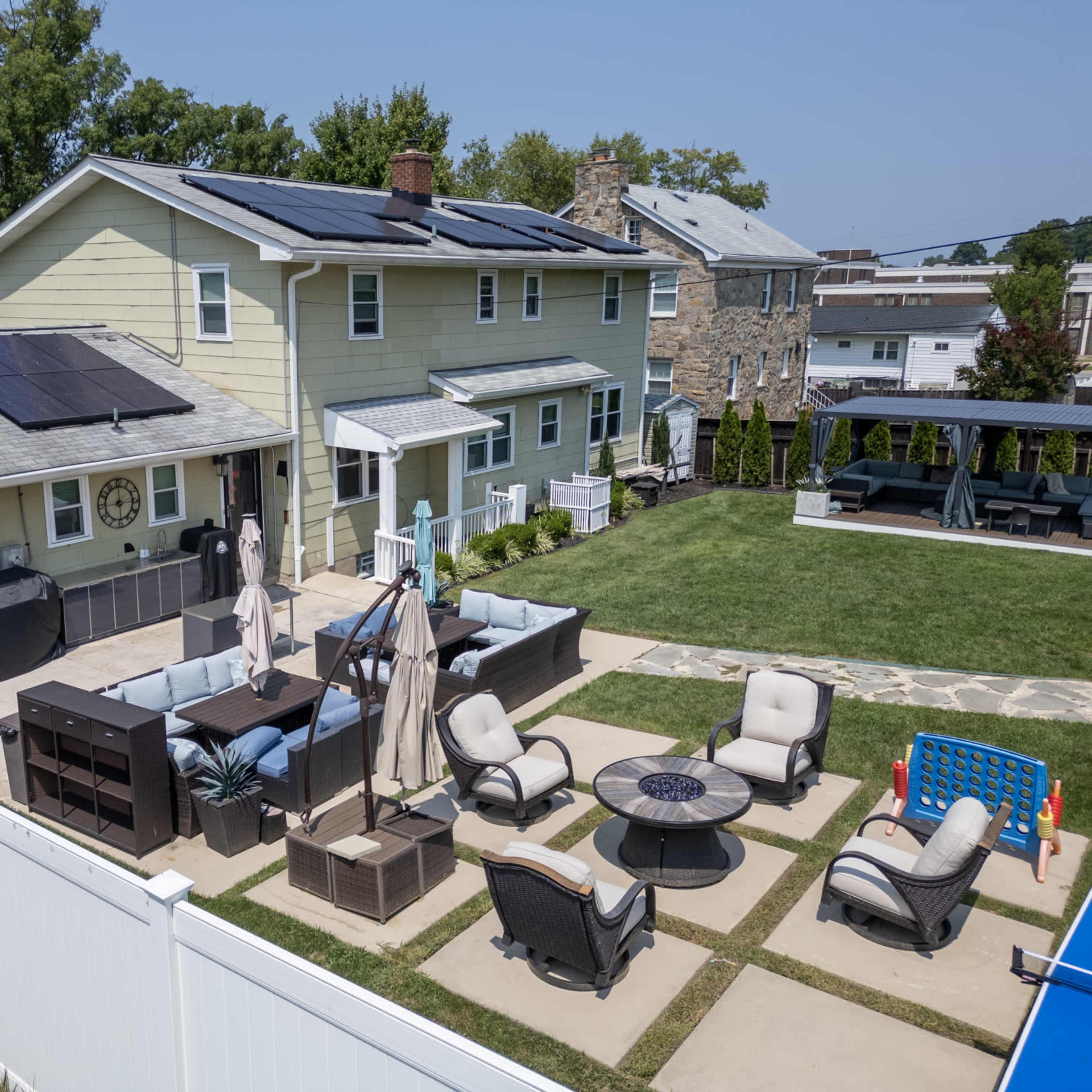 A backyard scene features a two-story house with solar panels on the roof, surrounded by patio furniture and a grassy area, enclosed by a white fence.
