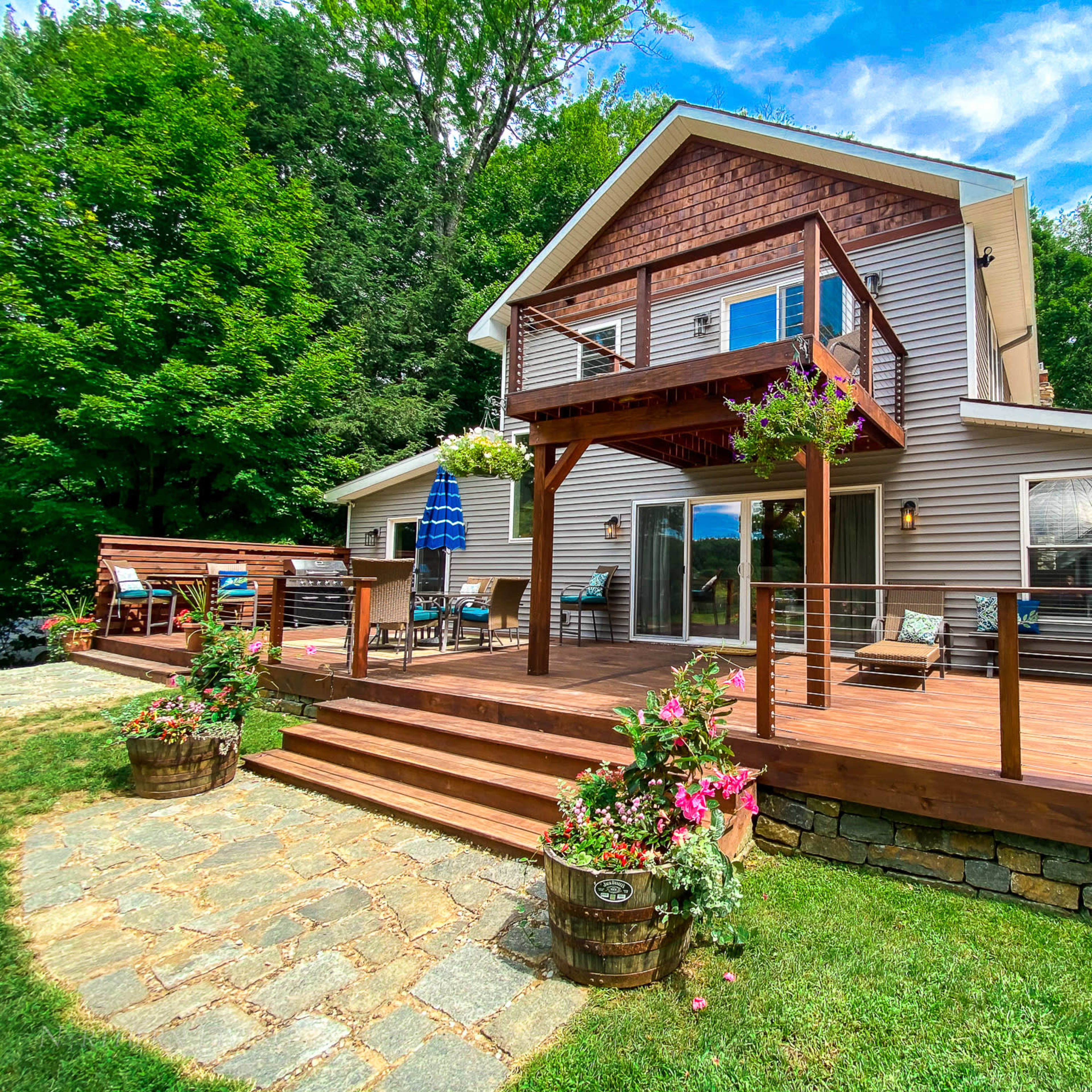 A two-story house with a wooden deck and a landscaped yard featuring stone pathways and flowerpots.
