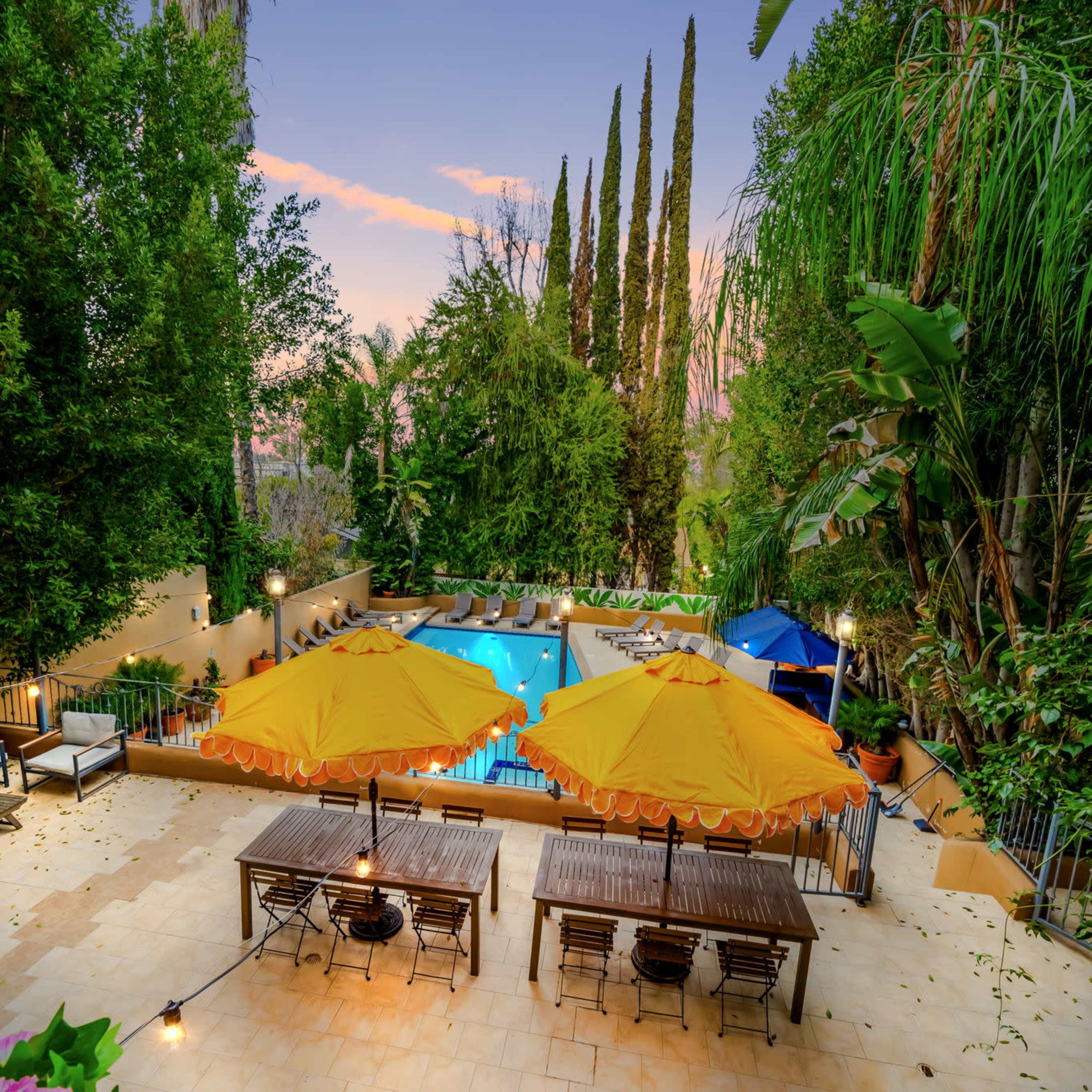 A backyard garden with a swimming pool, surrounded by tall trees, outdoor furniture, and two large orange umbrellas over a dining area.