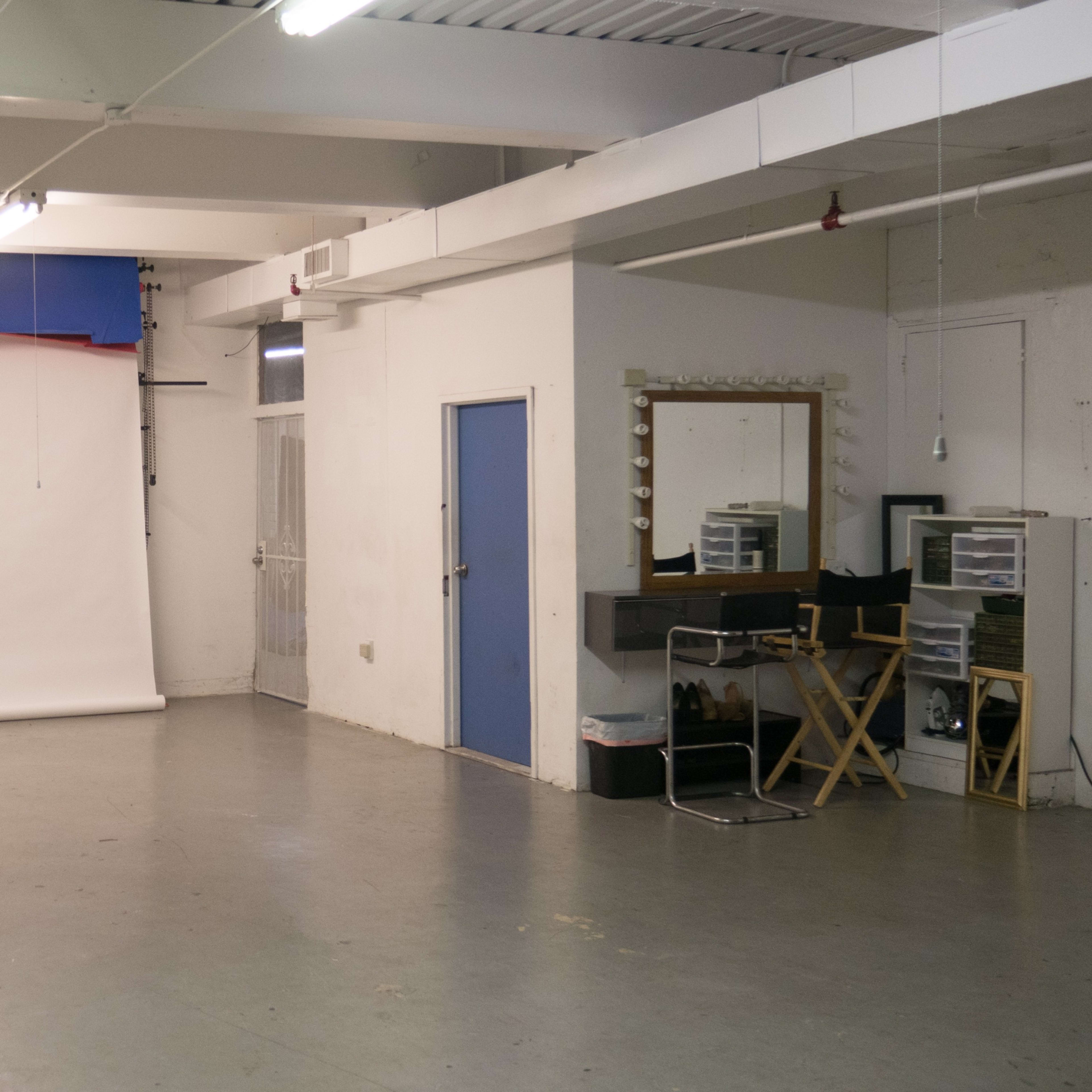 A sparsely furnished studio space with a white backdrop, a mirror, and a director's chair against a white brick wall.