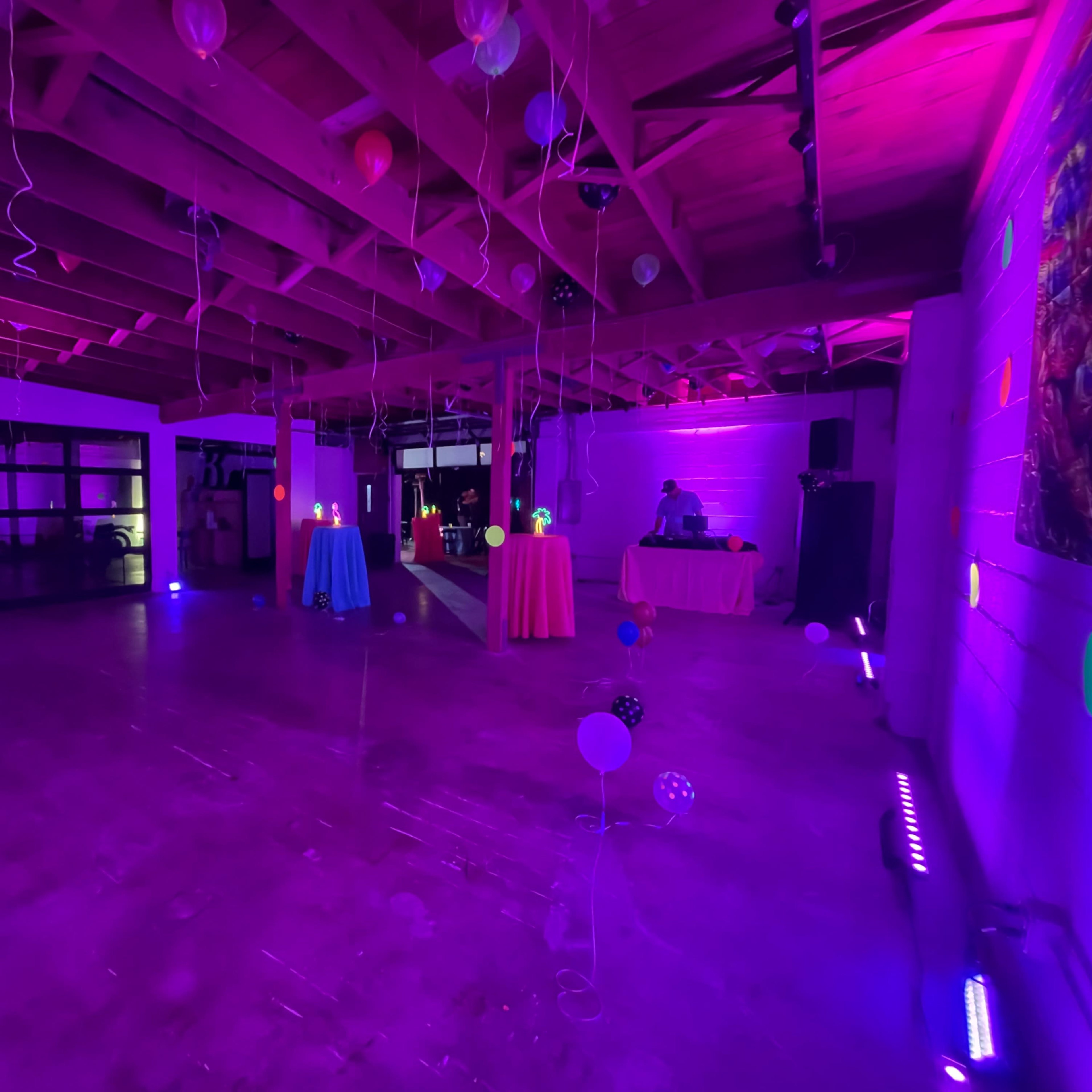The scene shows a spacious room with colorful tables covered in cloths, decorated with balloons and illuminated by purple lights.